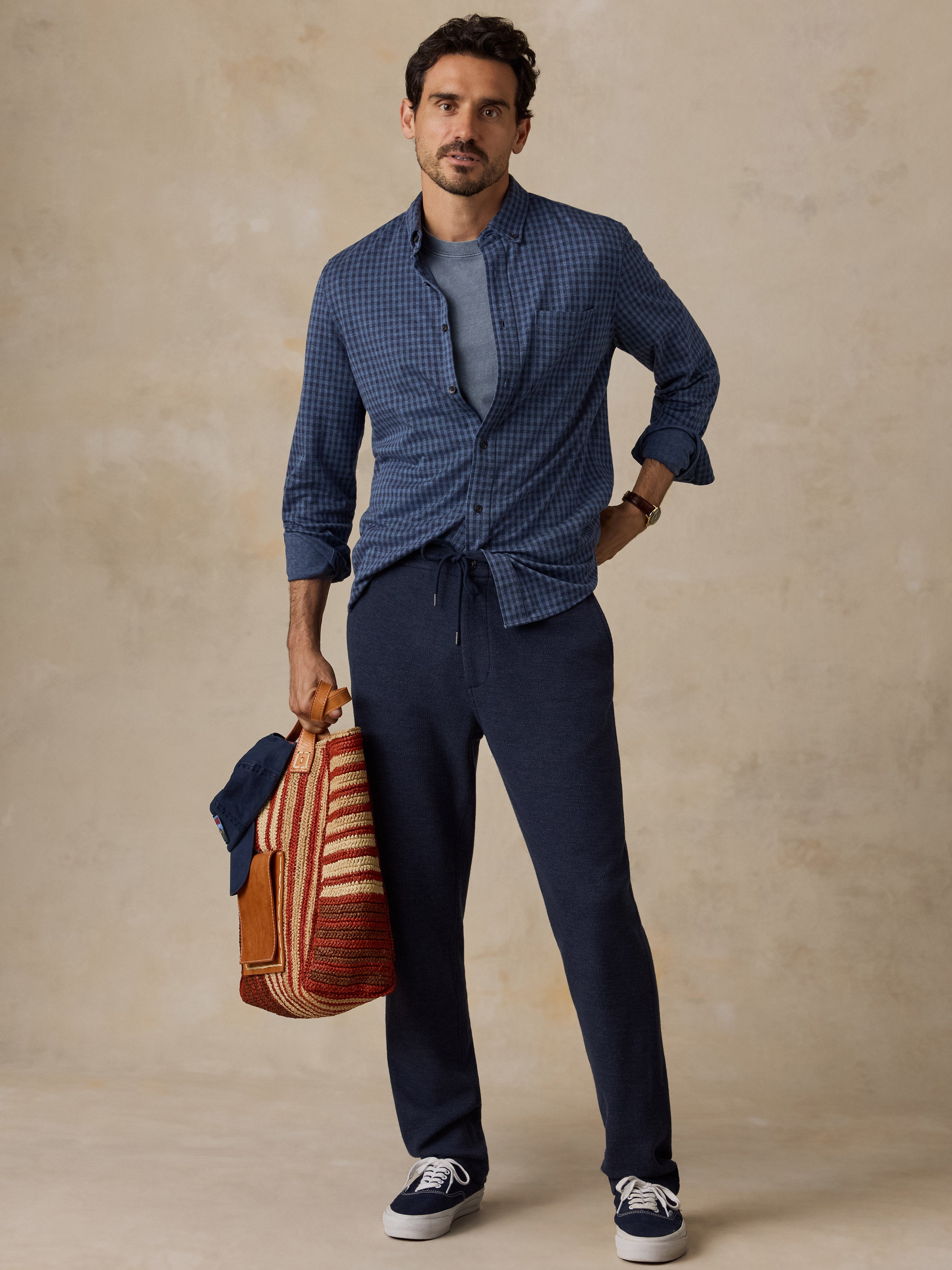 A man stands against a textured beige studio backdrop wearing a blue gingham button-up shirt over a gray T-shirt, navy drawstring trousers, and navy sneakers, while holding a striped woven bag.