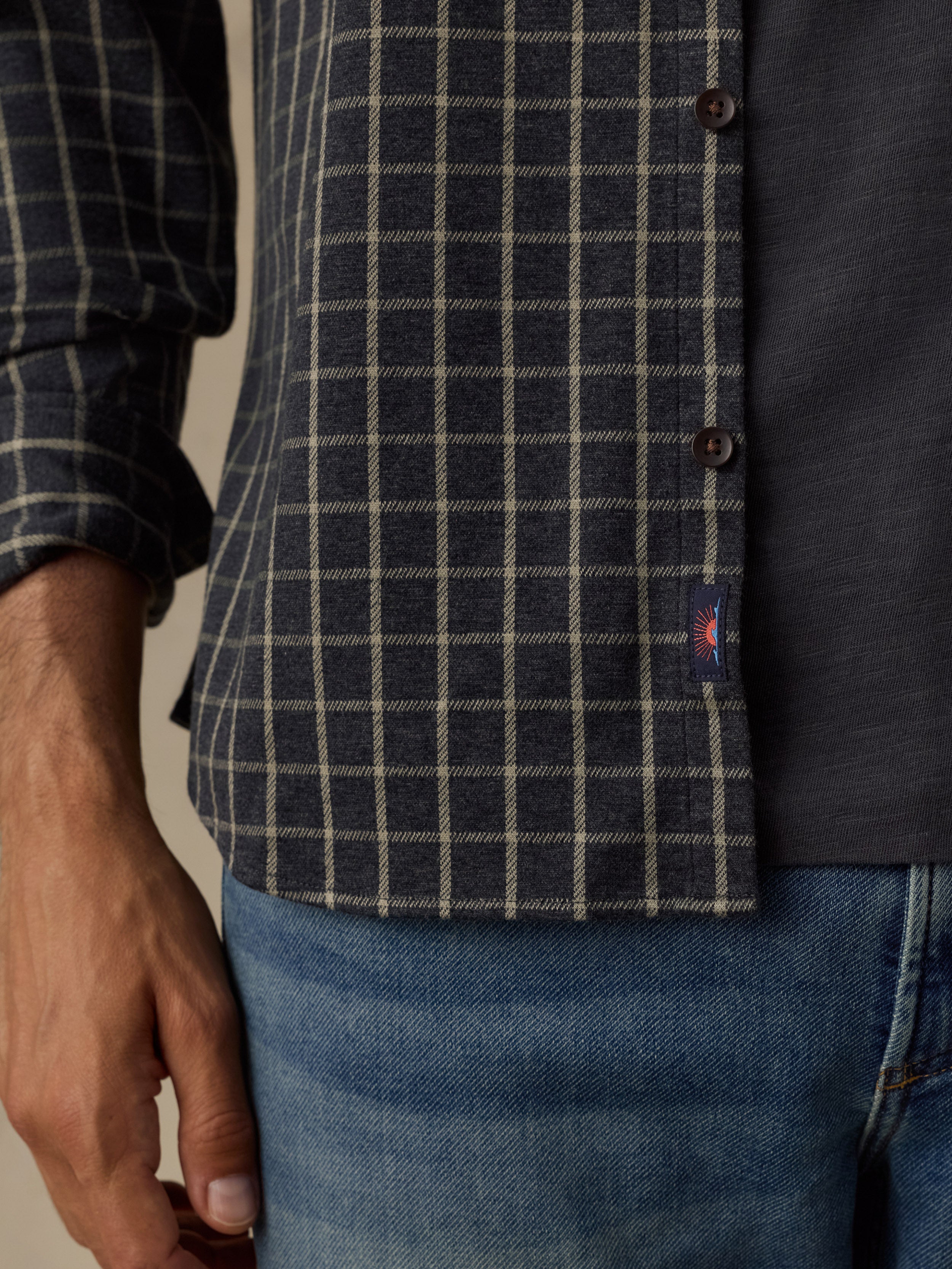 Close-up of a man wearing a navy and gray windowpane plaid button-down shirt with blue jeans, showing the shirt's texture, buttons, and a small red embroidered logo tab.