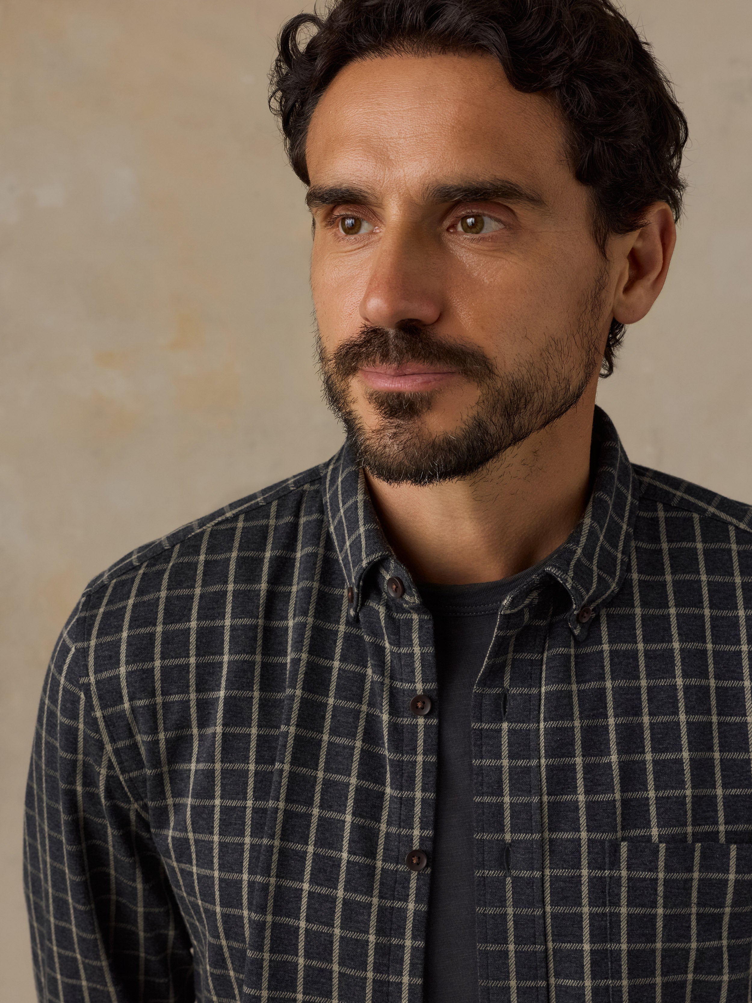 A man wearing a dark charcoal gray and black plaid button-down shirt over a black t-shirt, photographed against a textured beige background.