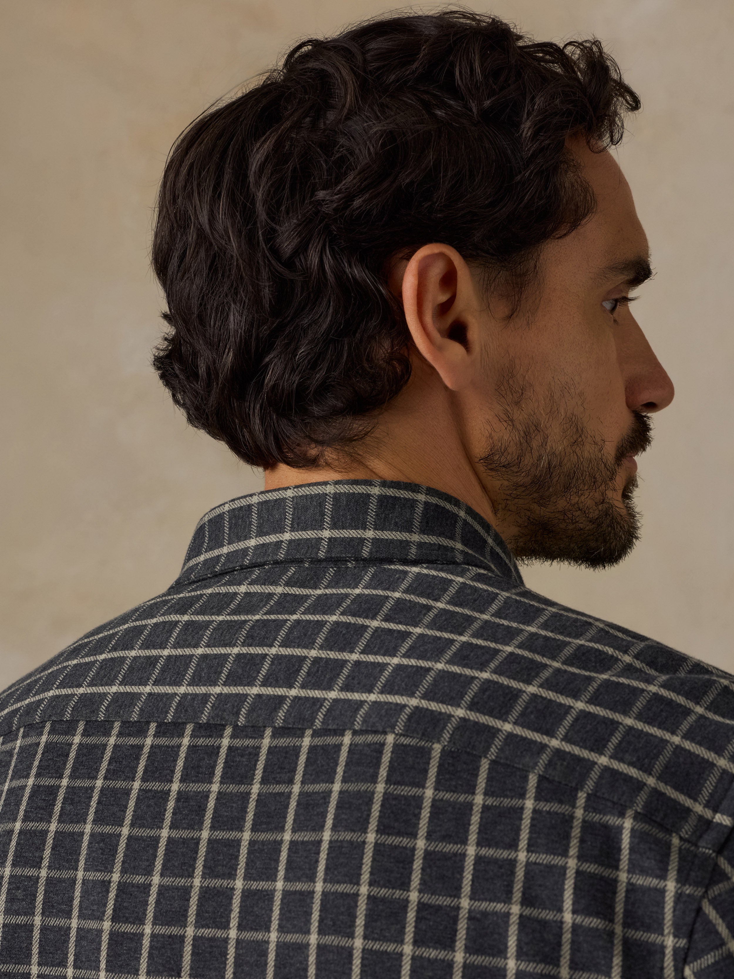 A man shown from the back and side profile wearing a dark navy and beige plaid button-down shirt against a neutral beige background.