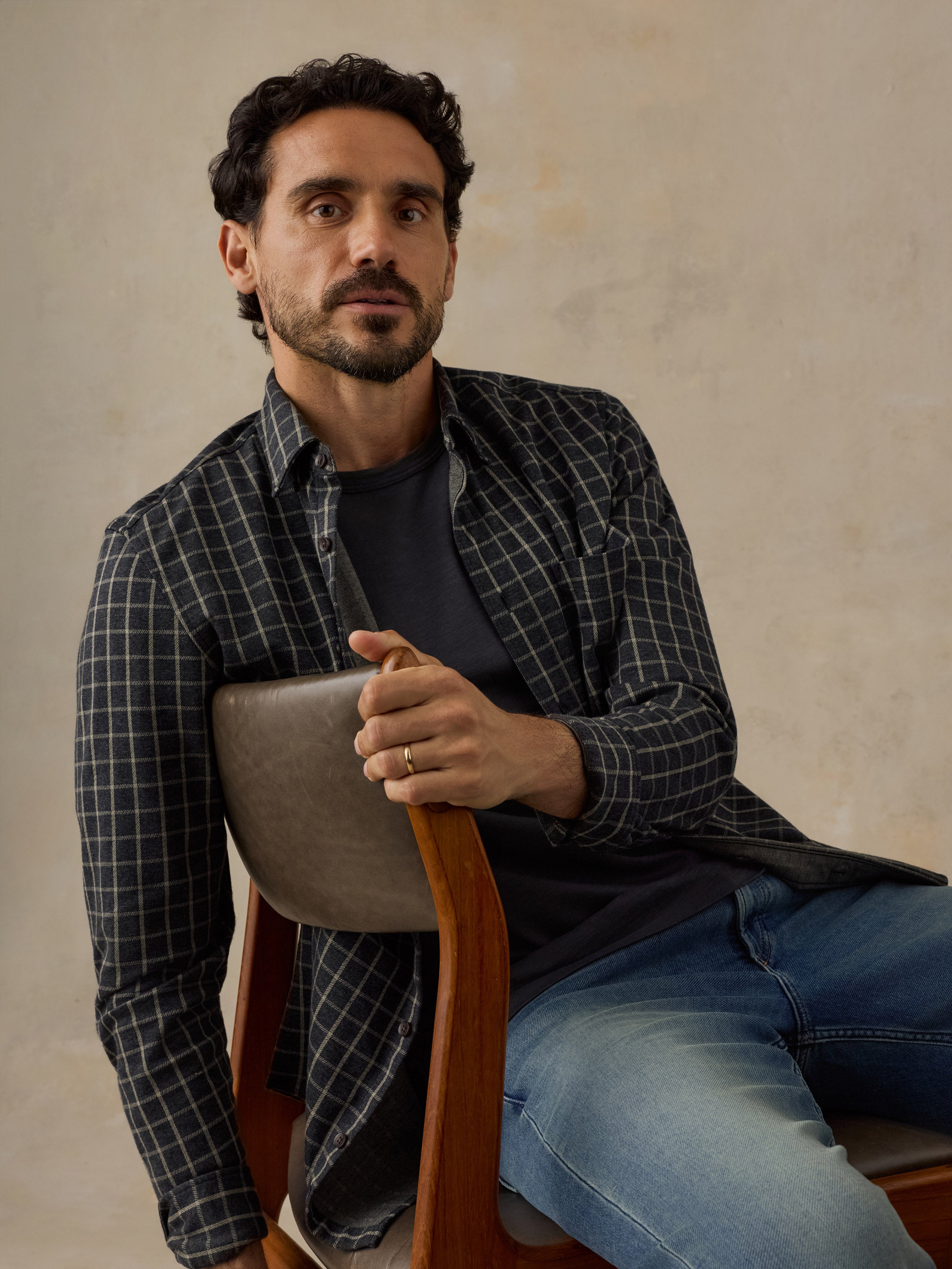 A man wearing a dark gray and black plaid button-down shirt over a black t-shirt and blue jeans, sitting on a wooden chair against a textured beige wall.