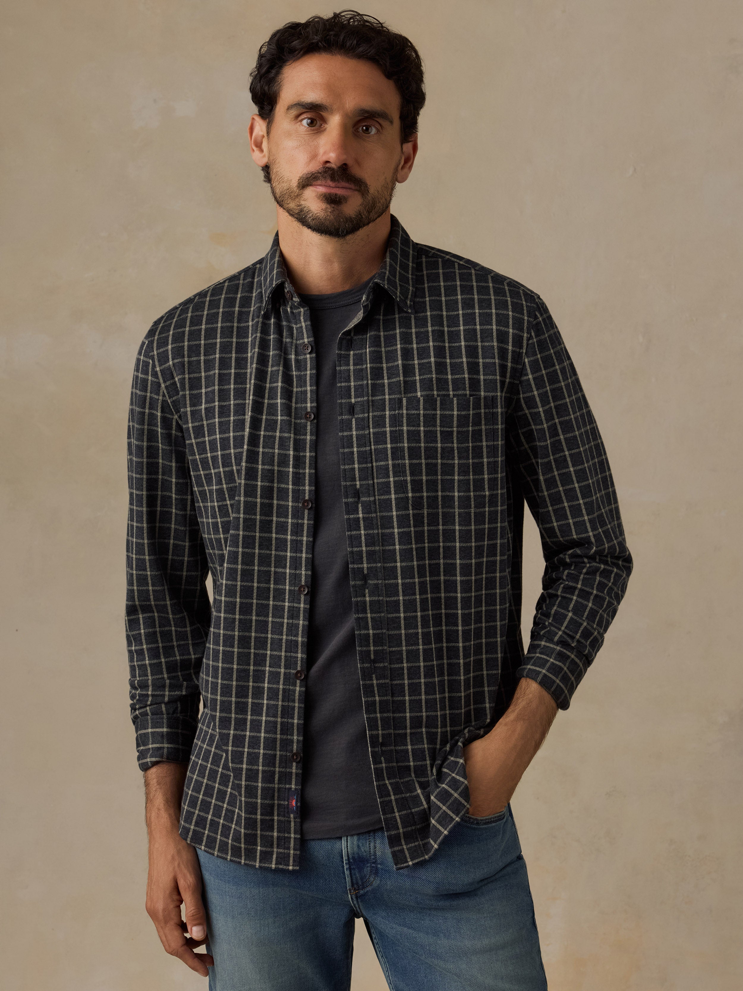 A man wearing a dark charcoal gray long-sleeve button-up shirt with a white windowpane plaid pattern, layered over a dark gray t-shirt and blue jeans, standing against a textured beige studio backdrop.