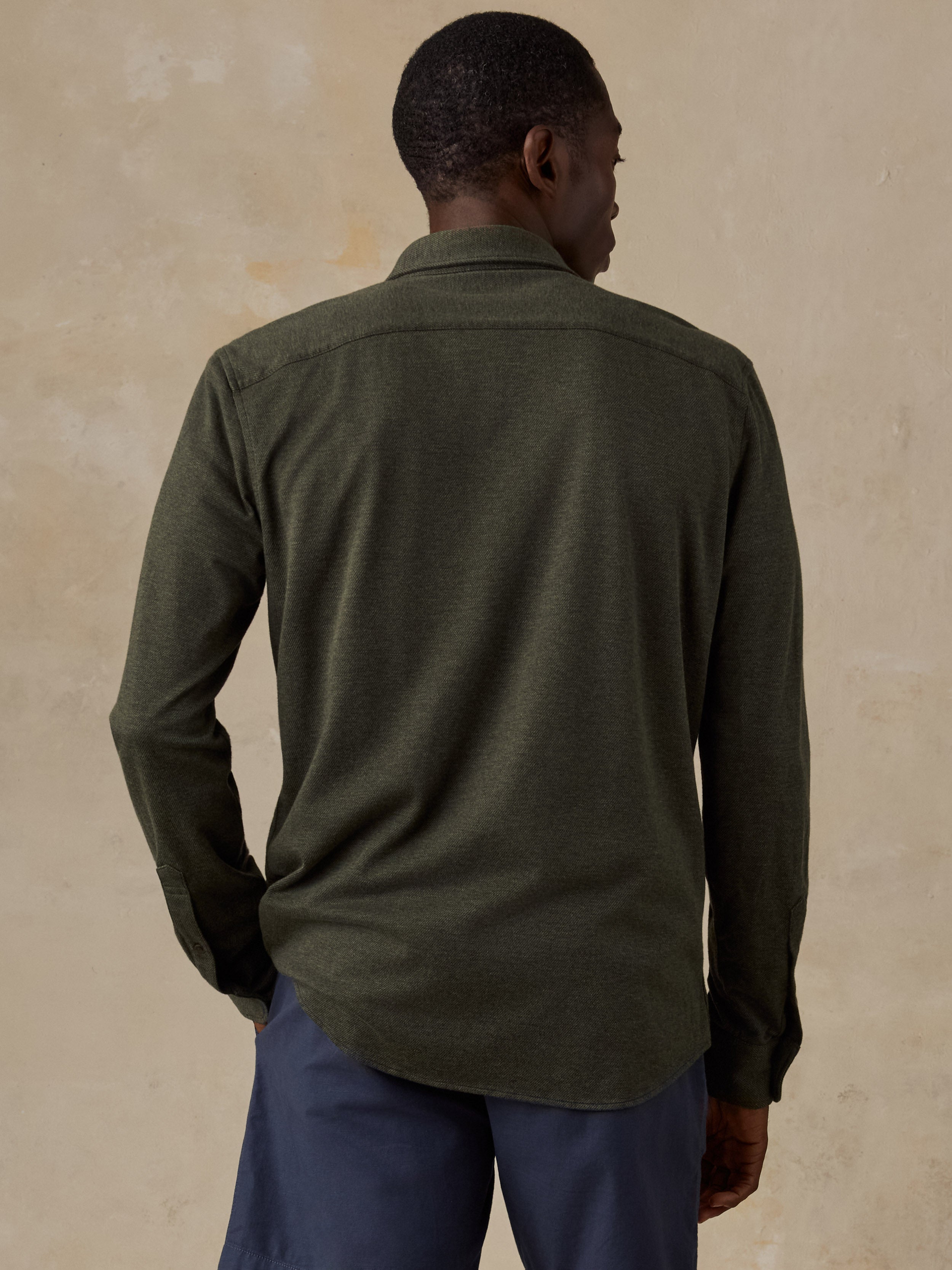 A man viewed from behind wearing an olive green long-sleeve button-up shirt and navy blue trousers, standing against a textured beige wall.