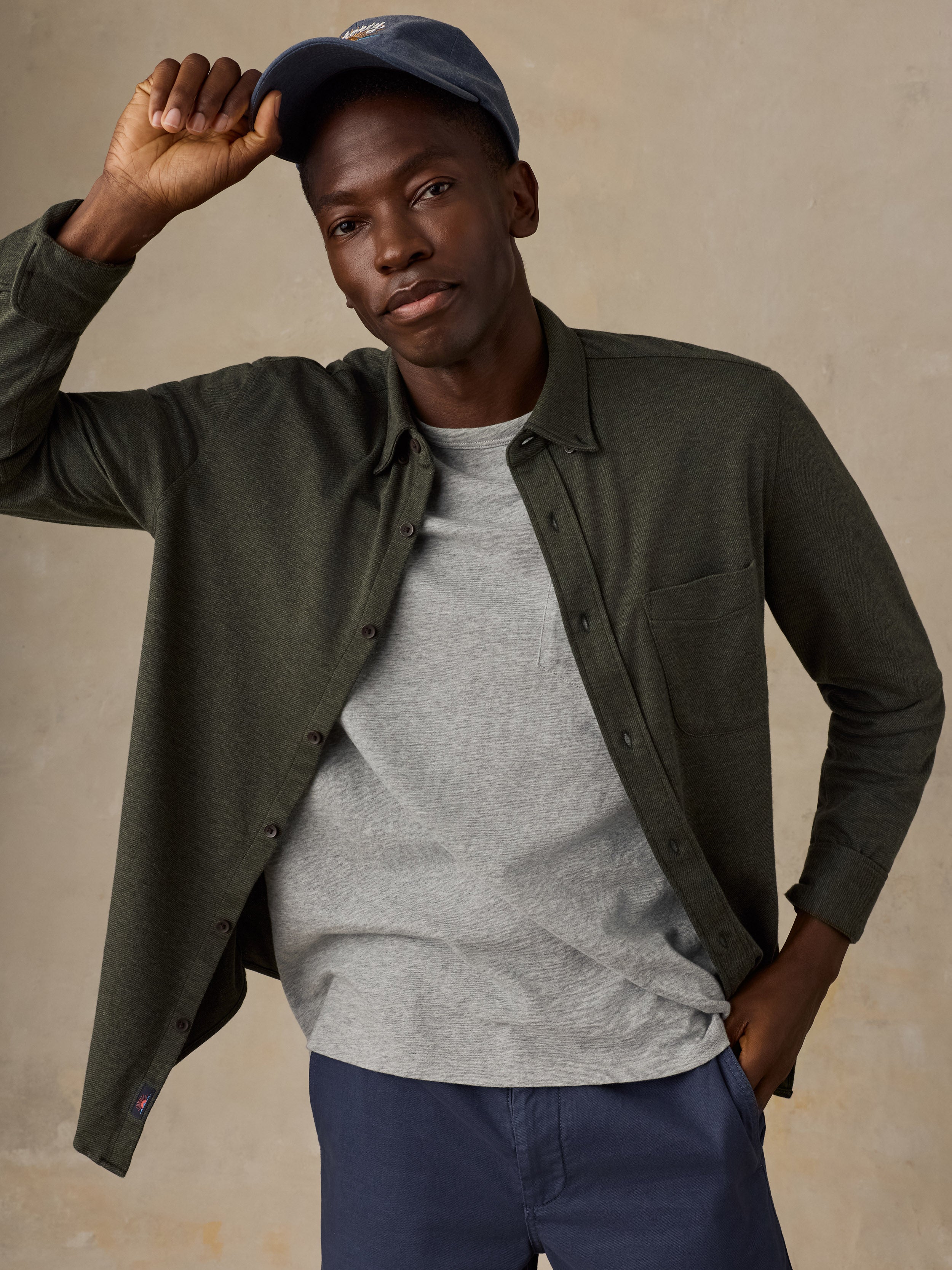 A man wearing an olive green long-sleeve button-up shirt over a heather gray t-shirt, paired with navy blue pants and a navy baseball cap, posing against a textured beige background.