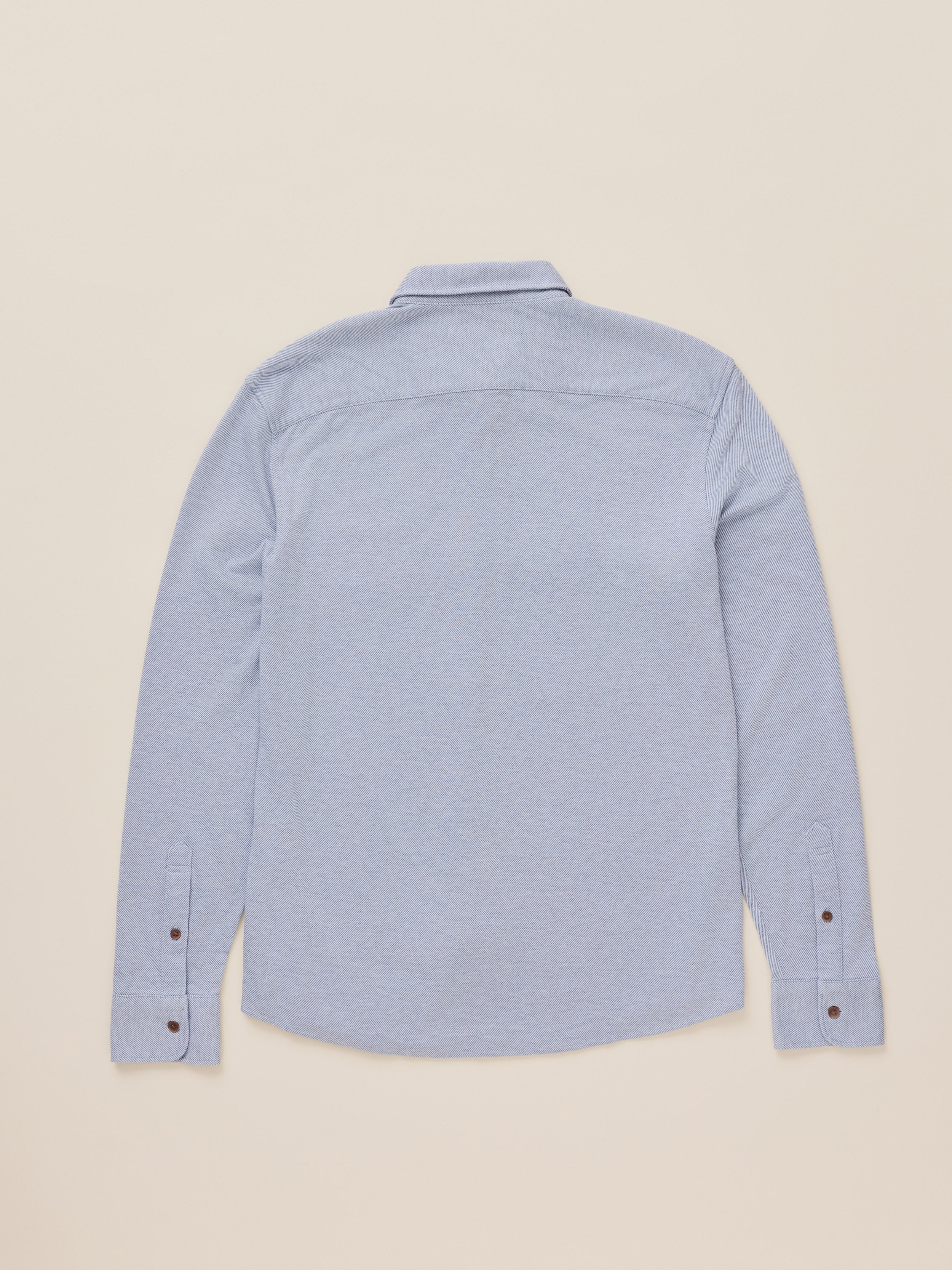 A light blue long-sleeve button-up shirt shown from the back, laid flat against a solid beige background.
