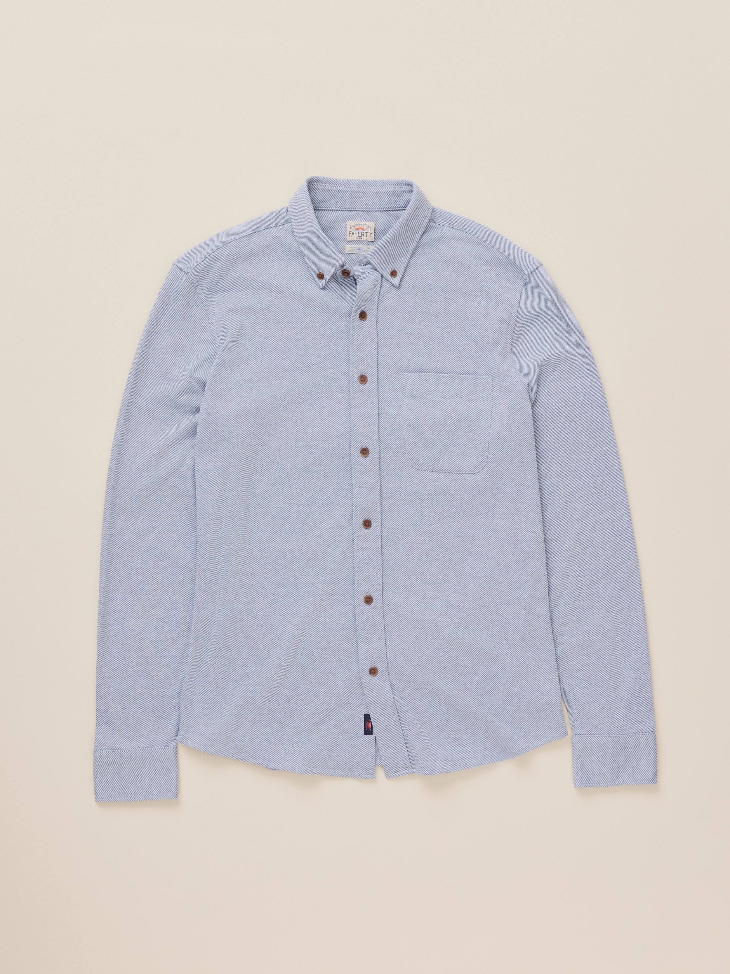 A light blue long-sleeve button-down shirt with a pointed collar and brown buttons, laid flat against a solid beige background.