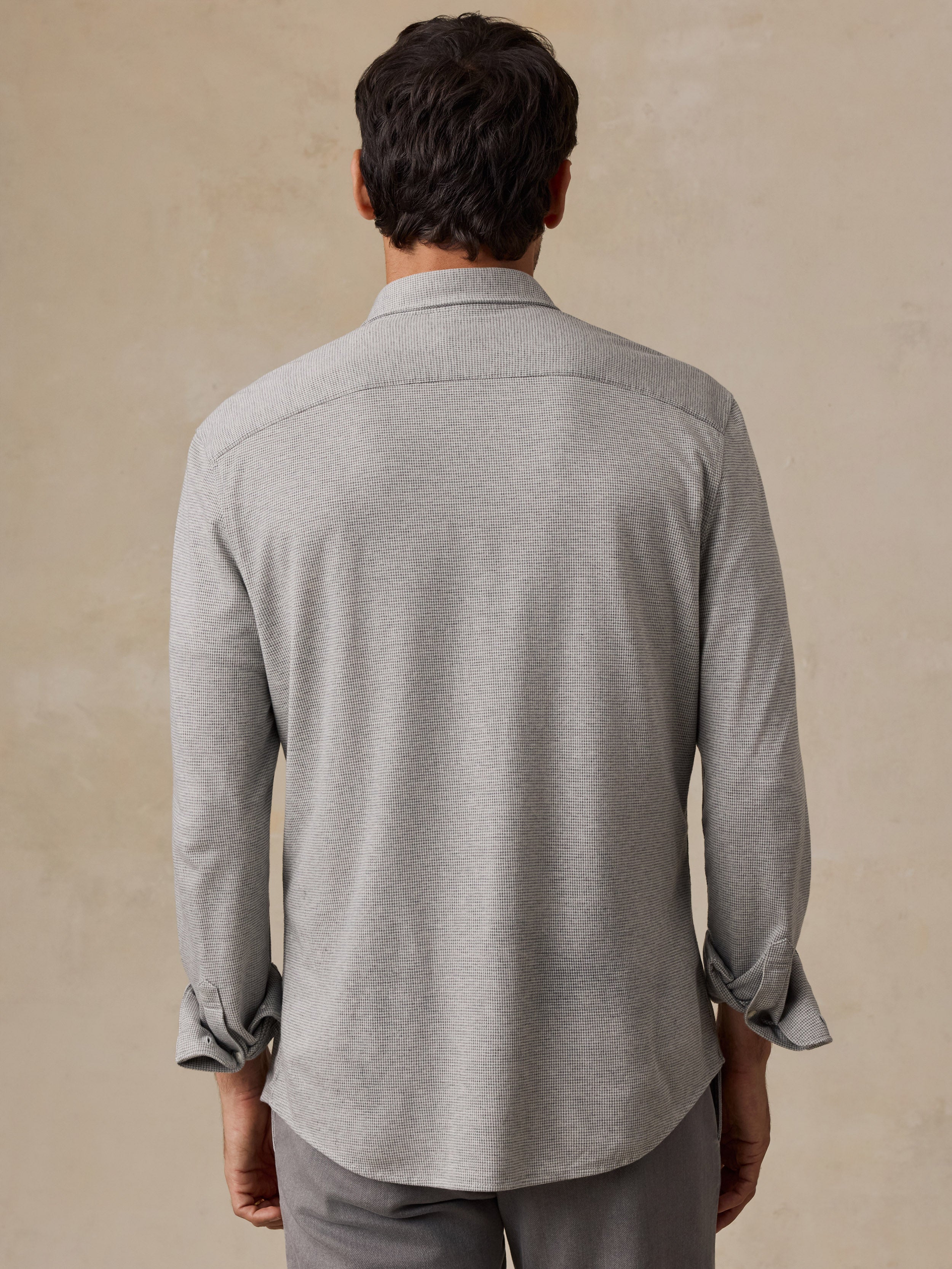 A man viewed from the back wearing a light gray heathered long-sleeve button-down shirt and gray trousers, standing against a textured beige background.