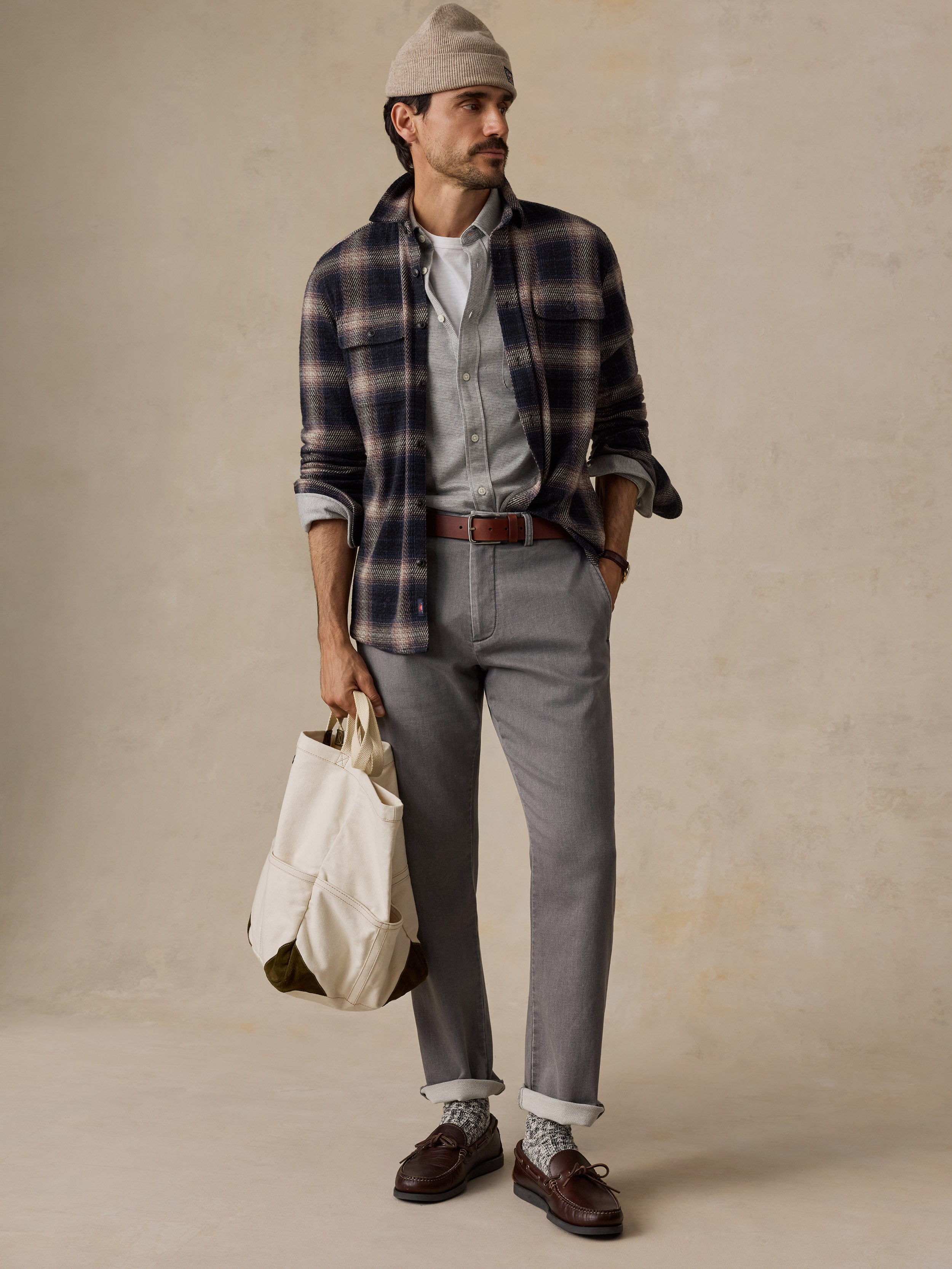 A man stands against a textured beige studio backdrop wearing a beige beanie, a navy and brown plaid flannel shirt over a light gray button-down, gray trousers, a brown belt, and dark brown loafers. He holds a cream canvas tote bag.