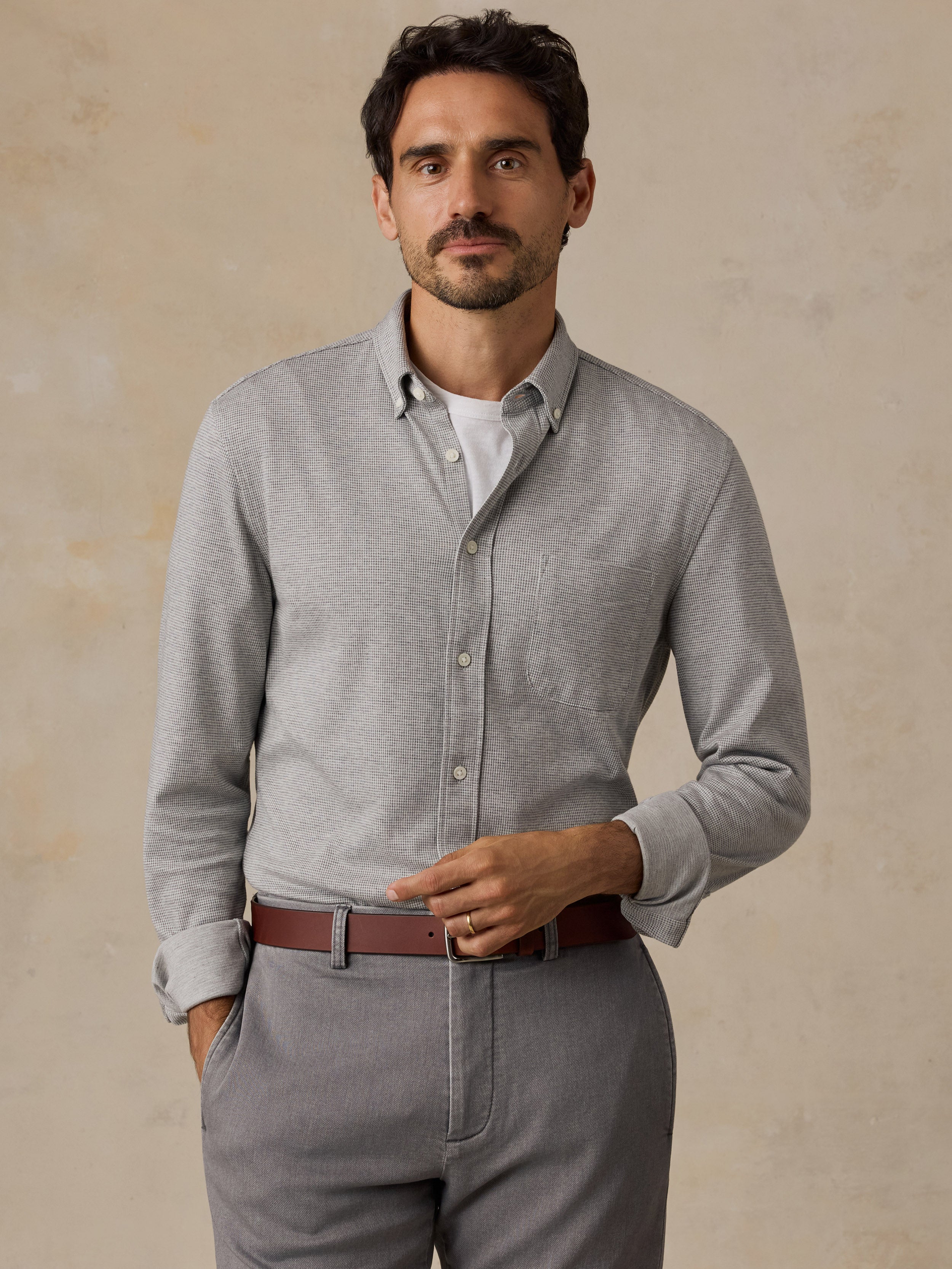 A man wearing a light gray long-sleeve knit button-down shirt over a white undershirt, paired with gray trousers and a brown belt, standing against a textured beige background.