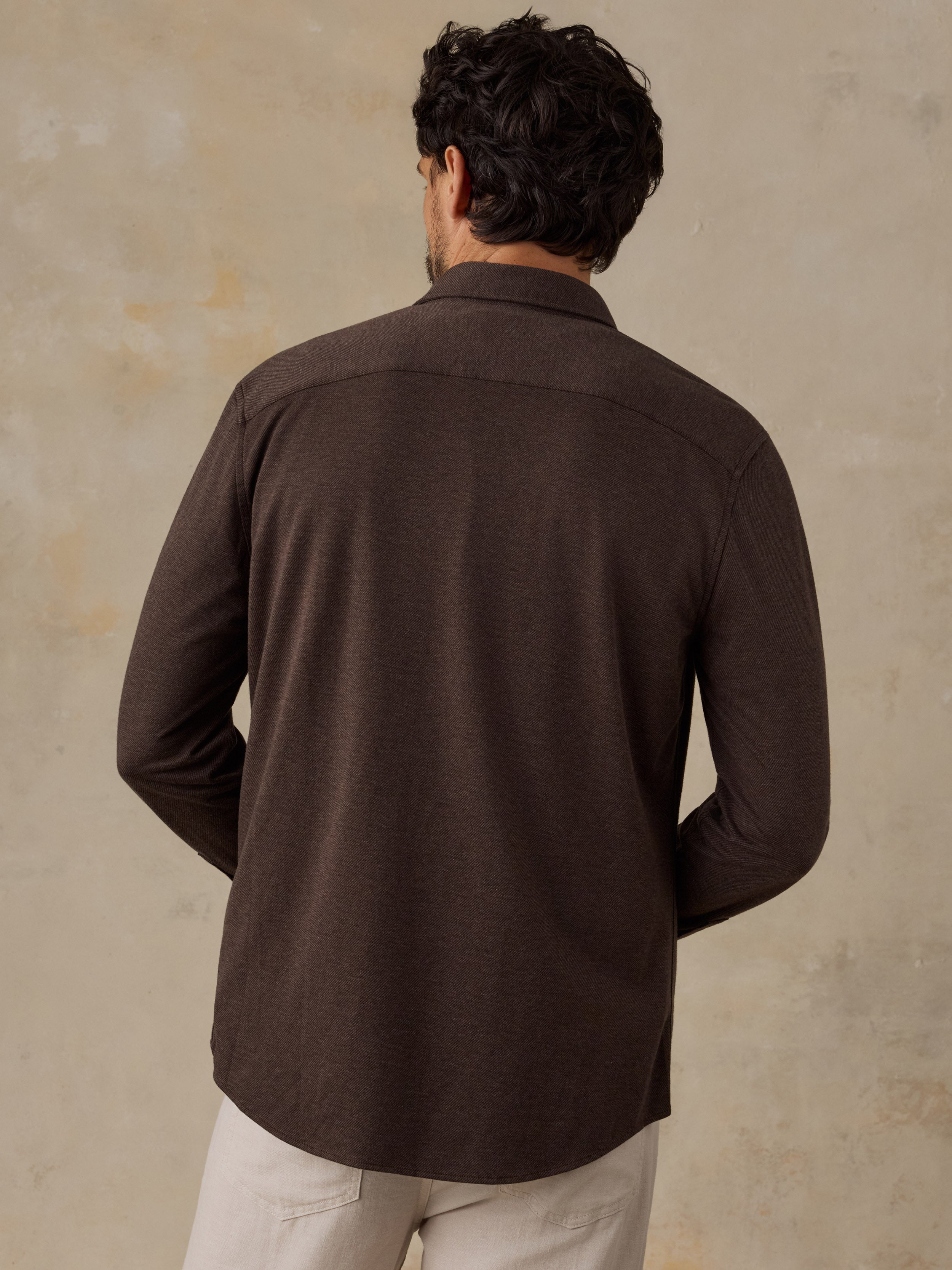 A man viewed from behind wearing a dark brown long-sleeve button-up shirt and light beige trousers, standing against a textured beige wall.