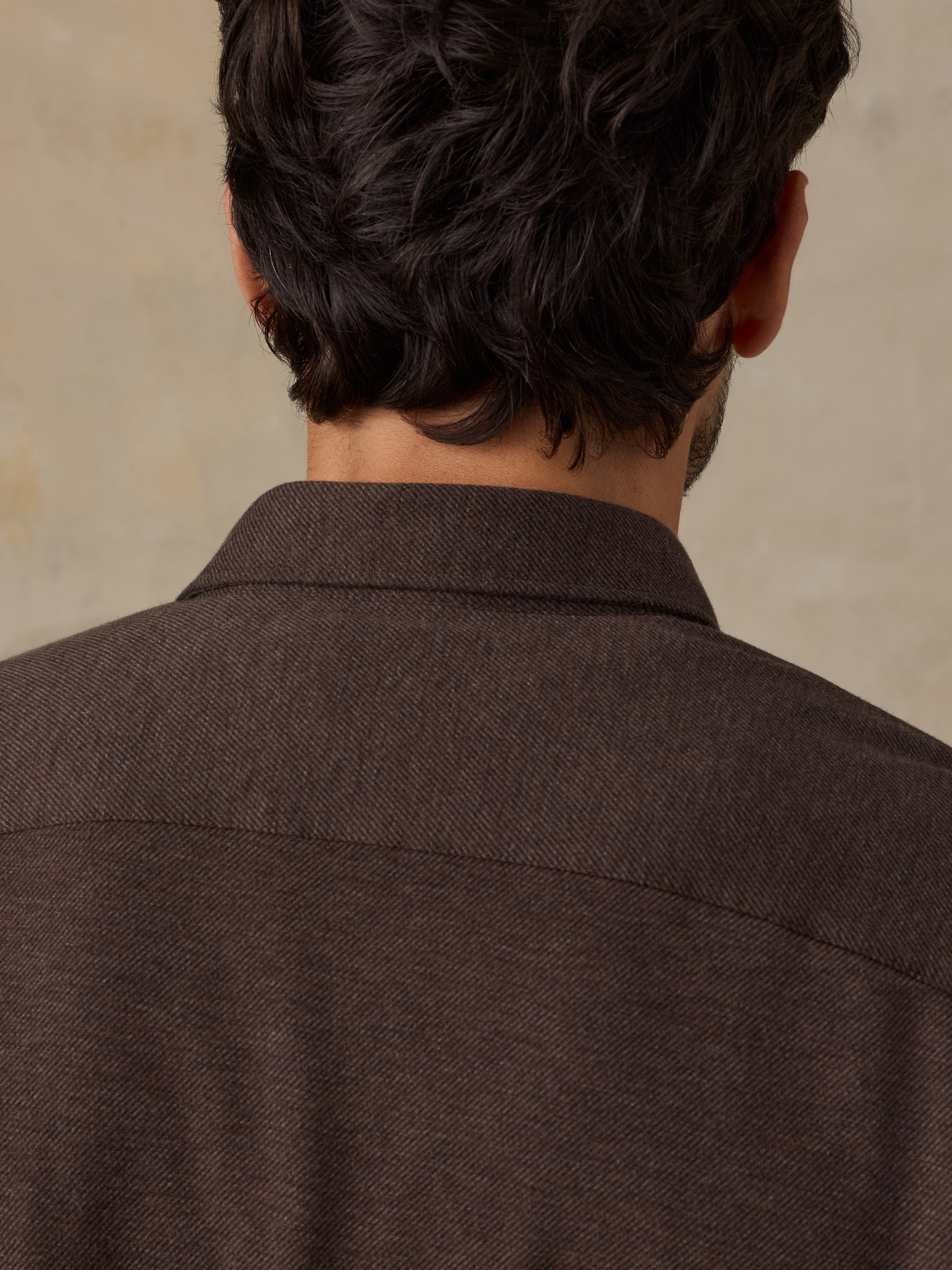 Close-up back view of a man wearing a dark brown textured collared shirt or jacket against a warm beige background.