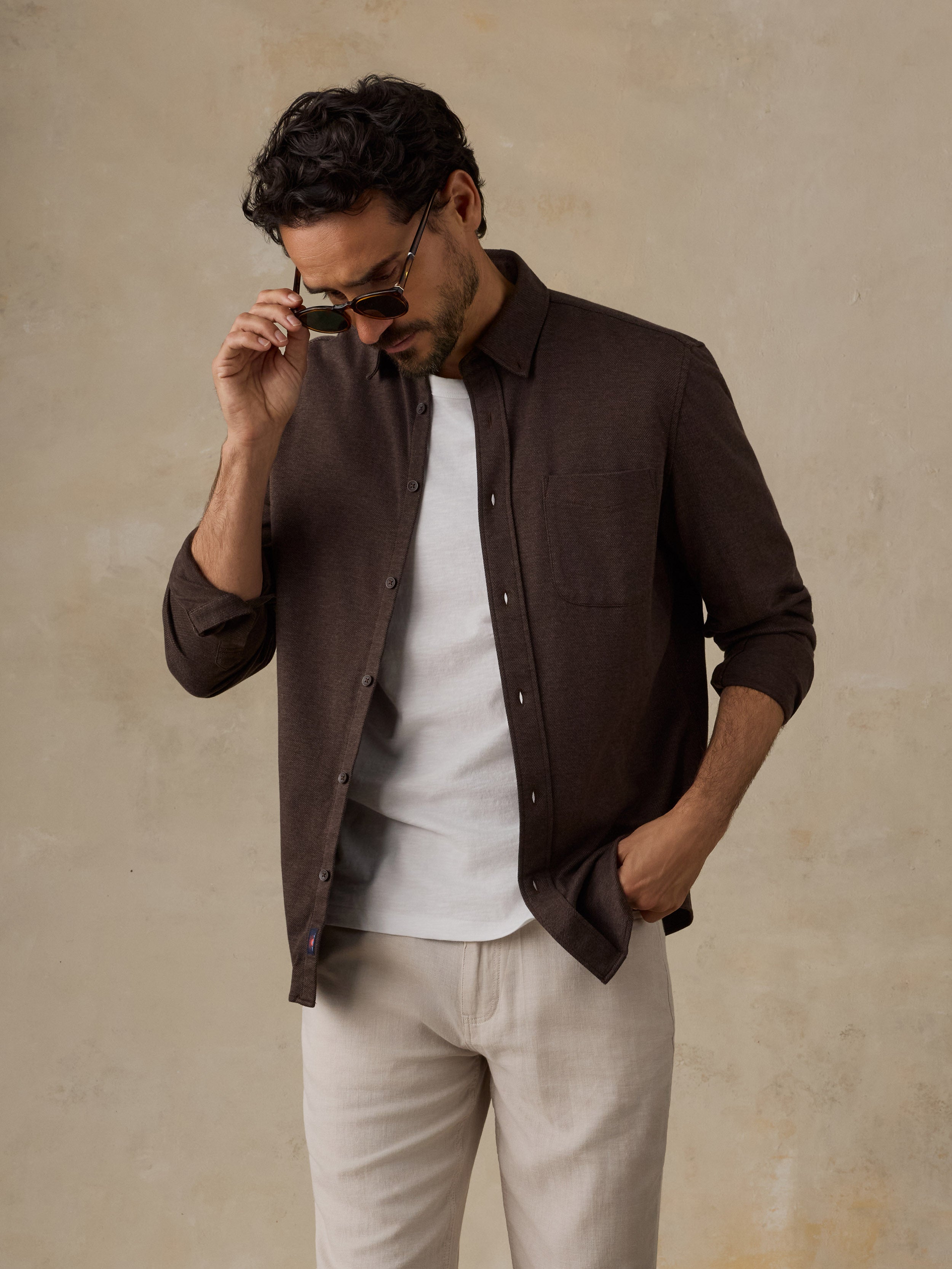 A man wearing a dark brown button-down shirt over a white t-shirt and beige trousers, standing against a textured beige wall.