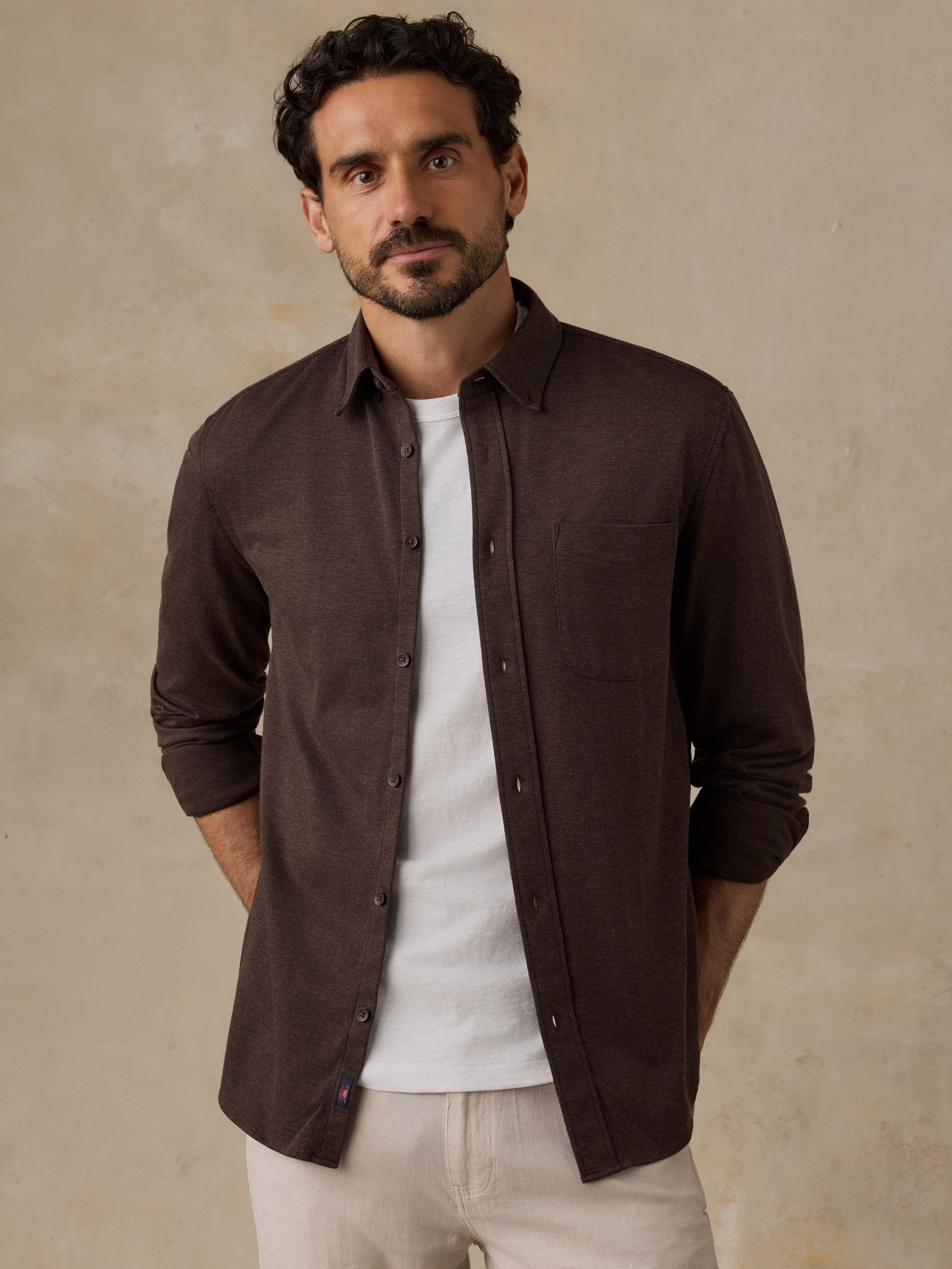A man wearing a dark brown long-sleeve button-down shirt over a white t-shirt and off-white trousers, standing against a textured beige wall.