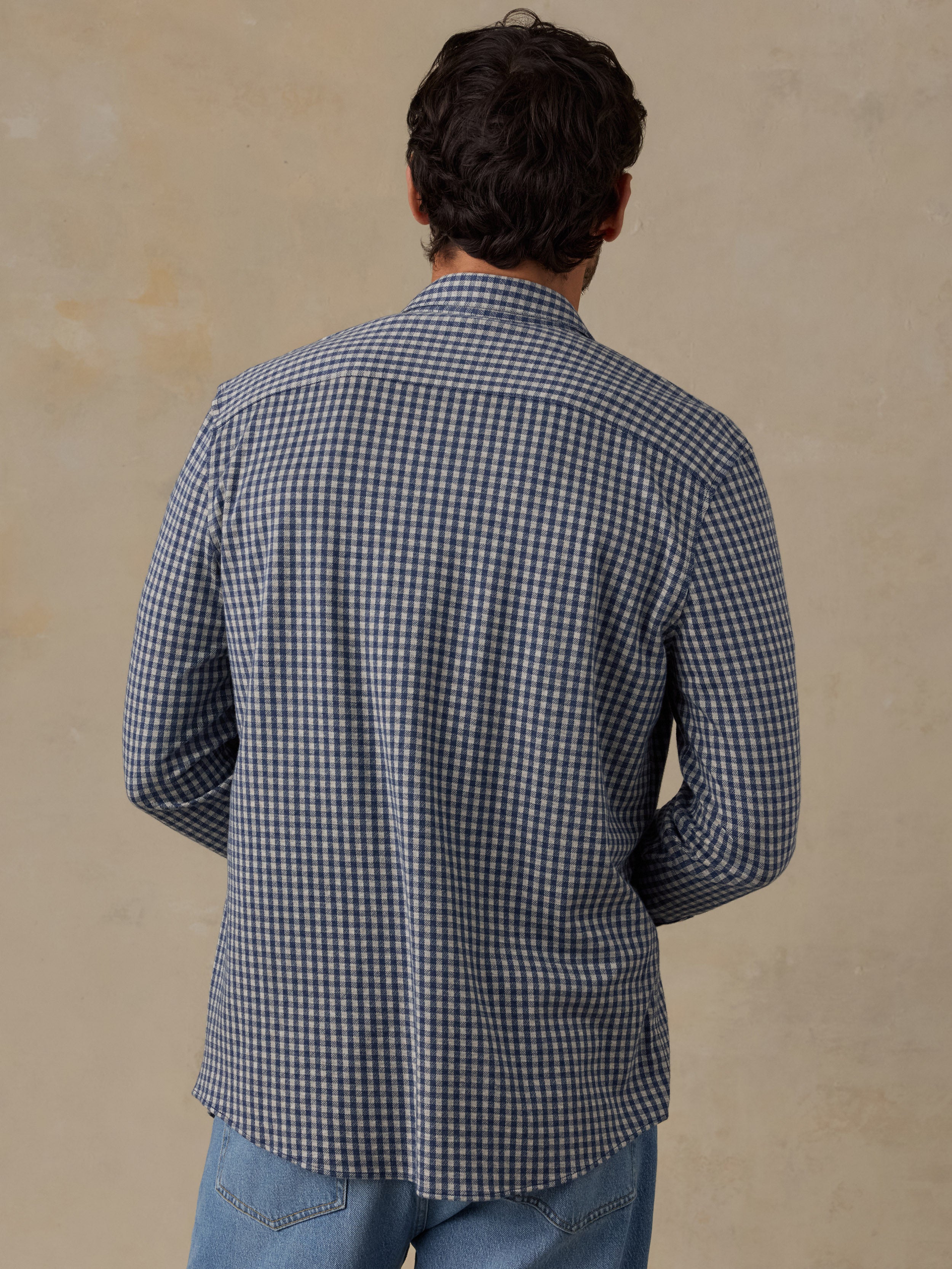 A man viewed from behind wearing a blue and white gingham long-sleeve button-down shirt and light blue jeans, standing against a textured beige wall.