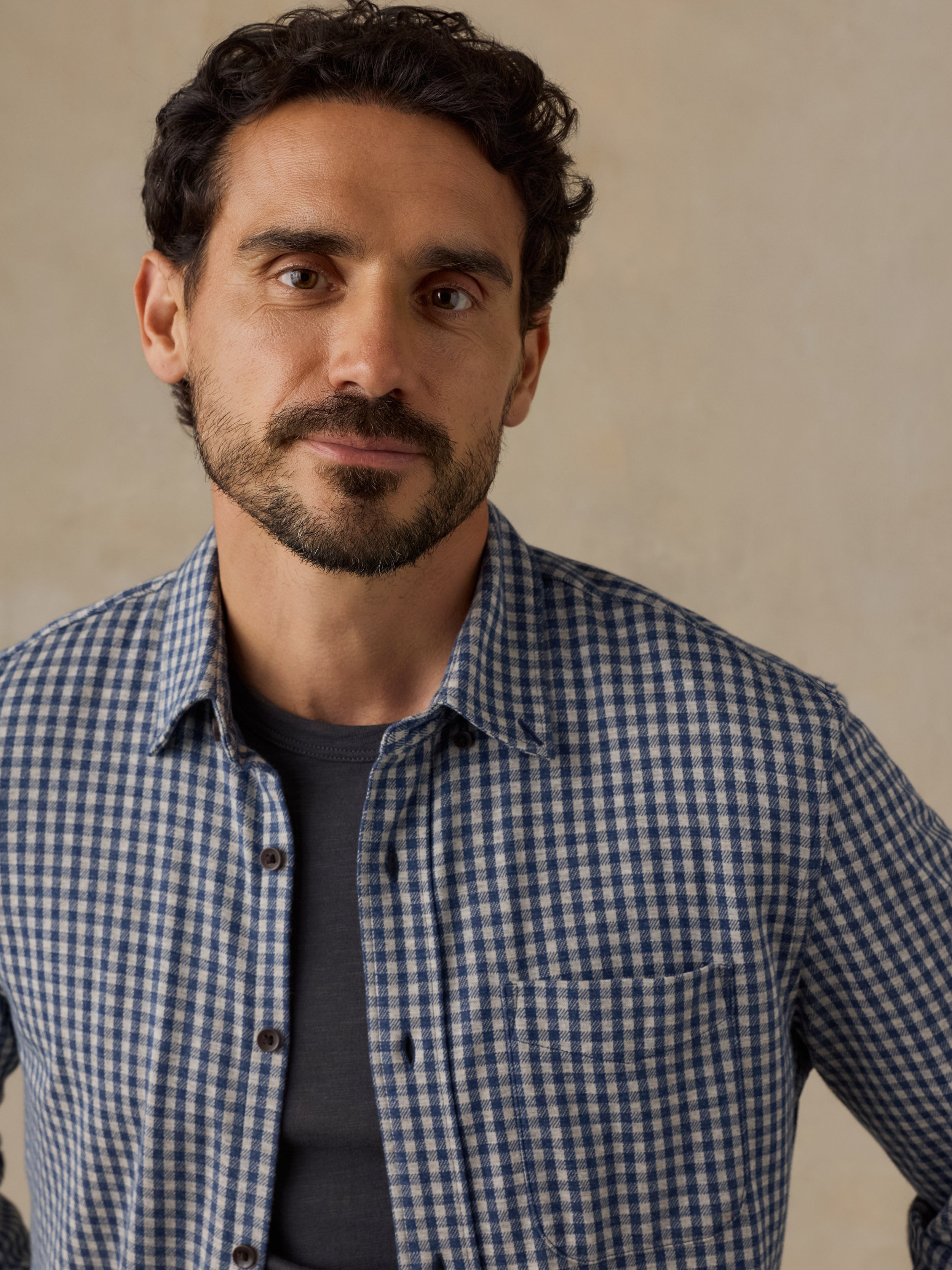 A man wearing a blue and white gingham check long-sleeve button-down shirt over a dark gray t-shirt, photographed against a neutral beige background.