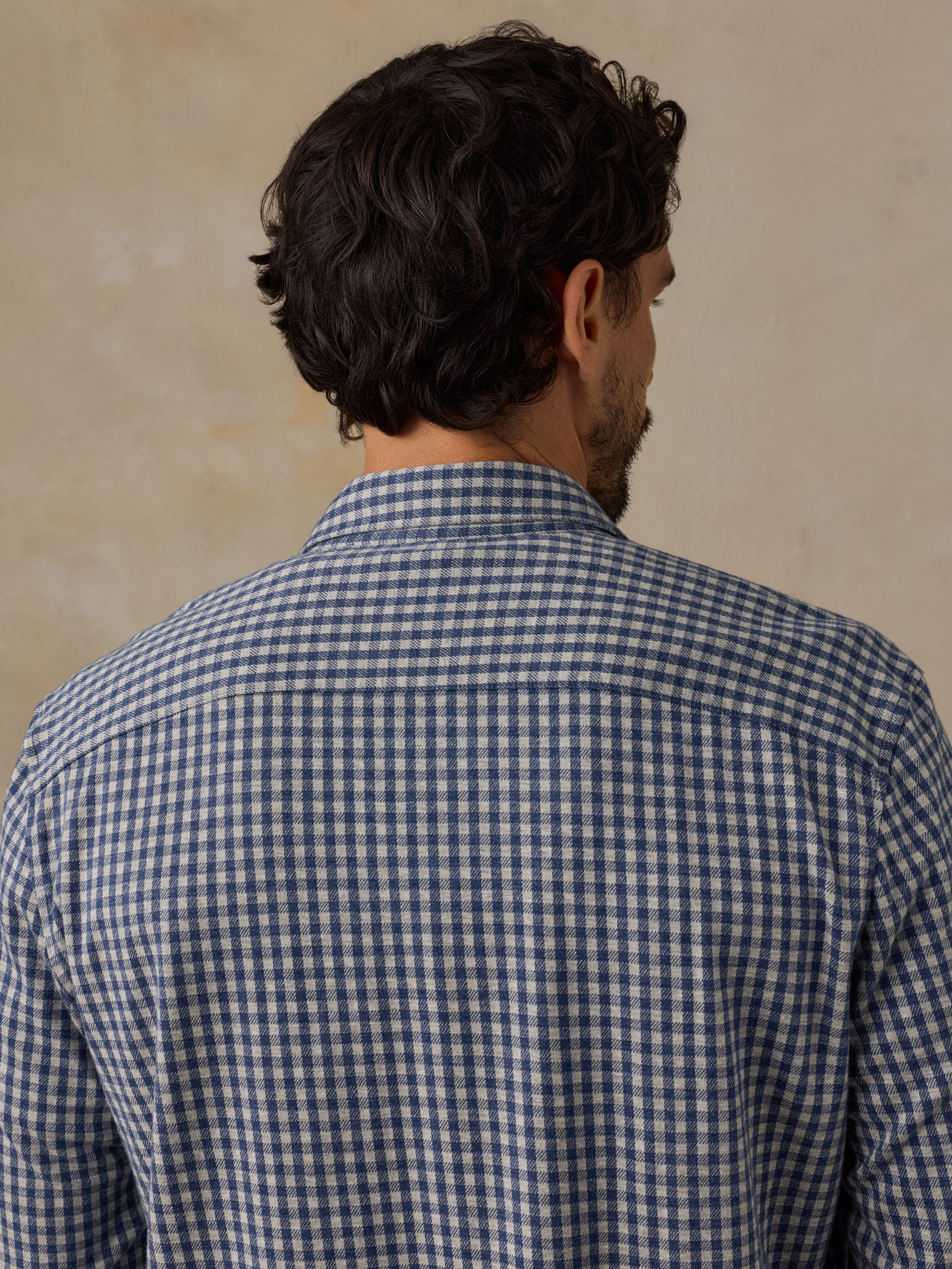 A man viewed from behind wearing a blue and white gingham check long-sleeve shirt against a textured beige background.
