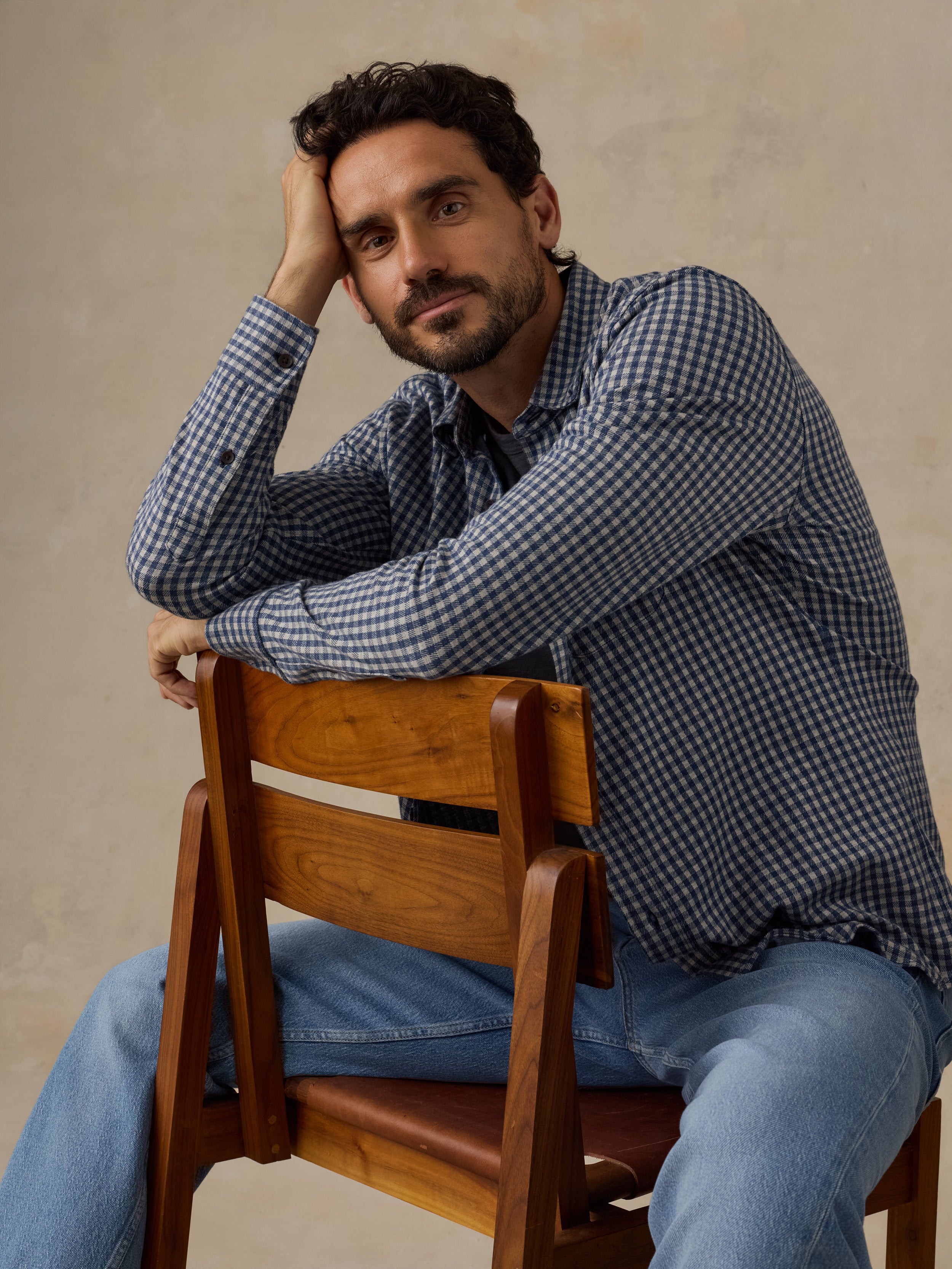 A man wearing a blue and white gingham long-sleeve button-up shirt and light blue jeans, sitting on a wooden chair against a textured beige wall.