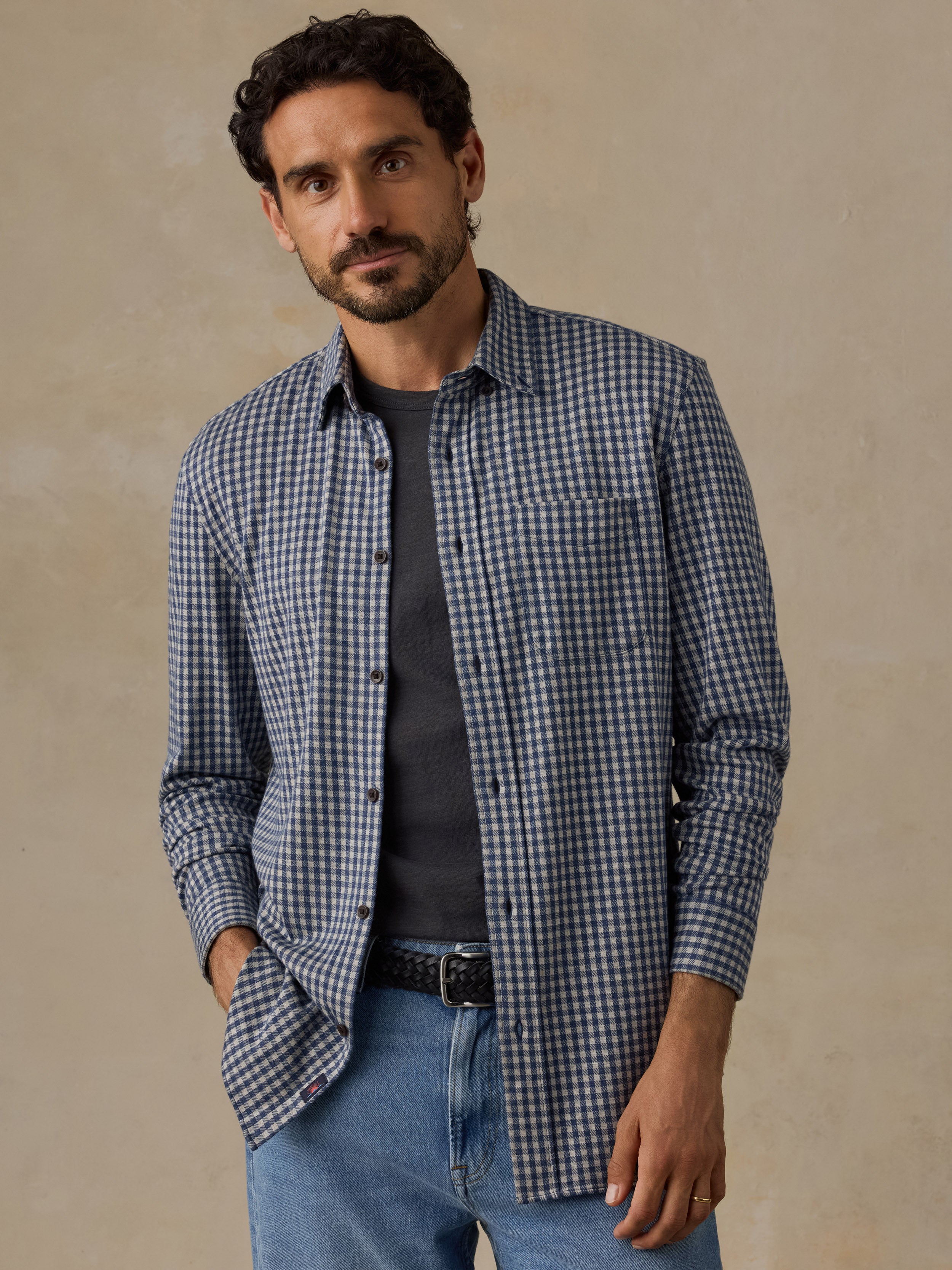 A man wearing a blue and white gingham check long-sleeve button-down shirt over a dark gray t-shirt, paired with blue jeans, standing against a textured beige background.