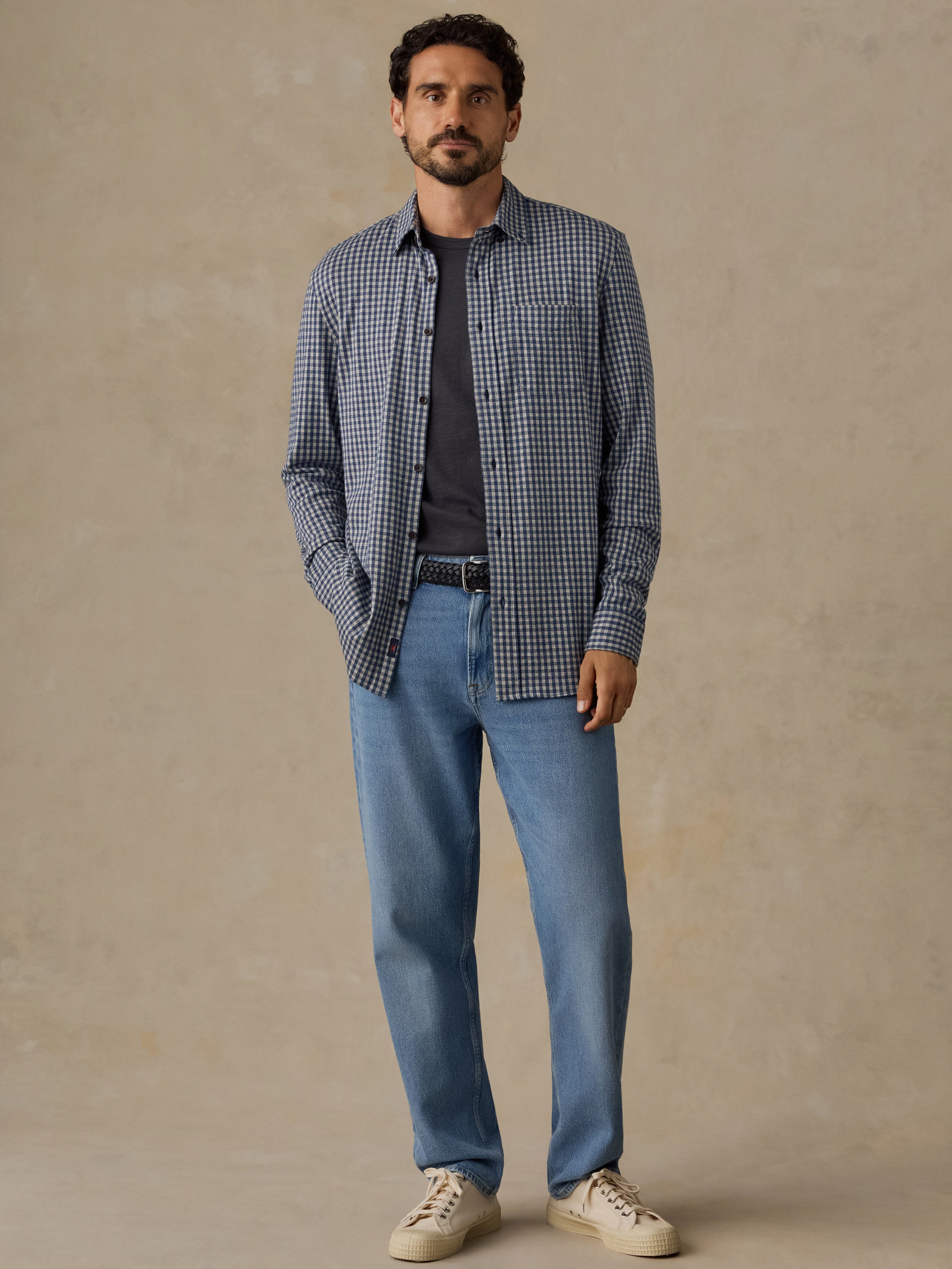 A man wearing a blue and white gingham check long-sleeve button-down shirt over a dark gray t-shirt, paired with light blue jeans and beige sneakers, standing against a textured beige studio backdrop.