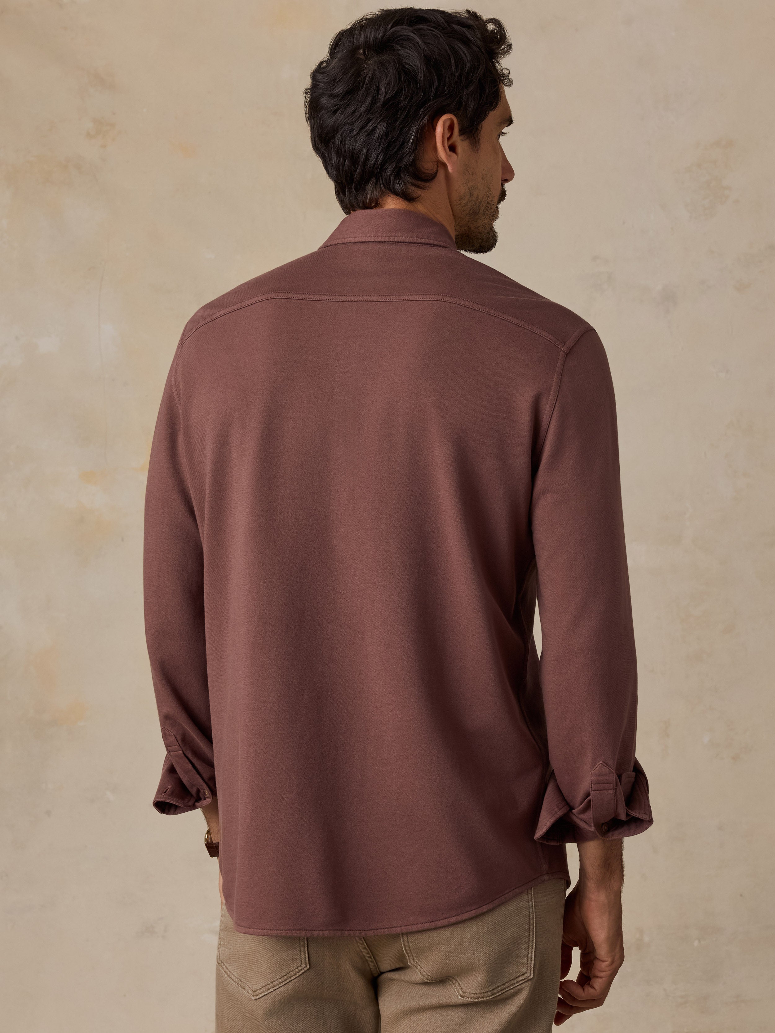A man shown from the back wearing a muted dusty rose long-sleeve button-up shirt and khaki pants, standing against a textured beige wall.