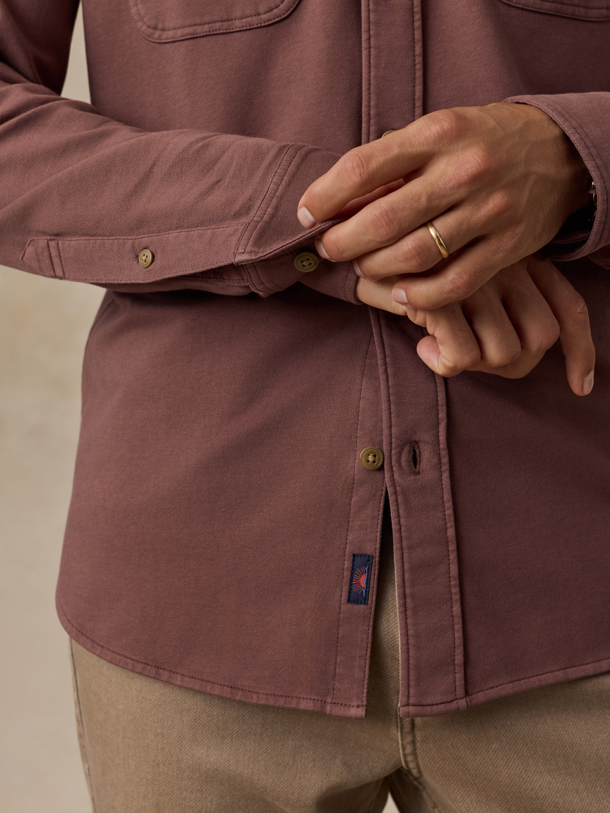 A close-up of a person wearing a dusty rose button-up shirt with tan pants, adjusting the cuff of the sleeve. A small blue and red woven brand tag is visible near the hem.