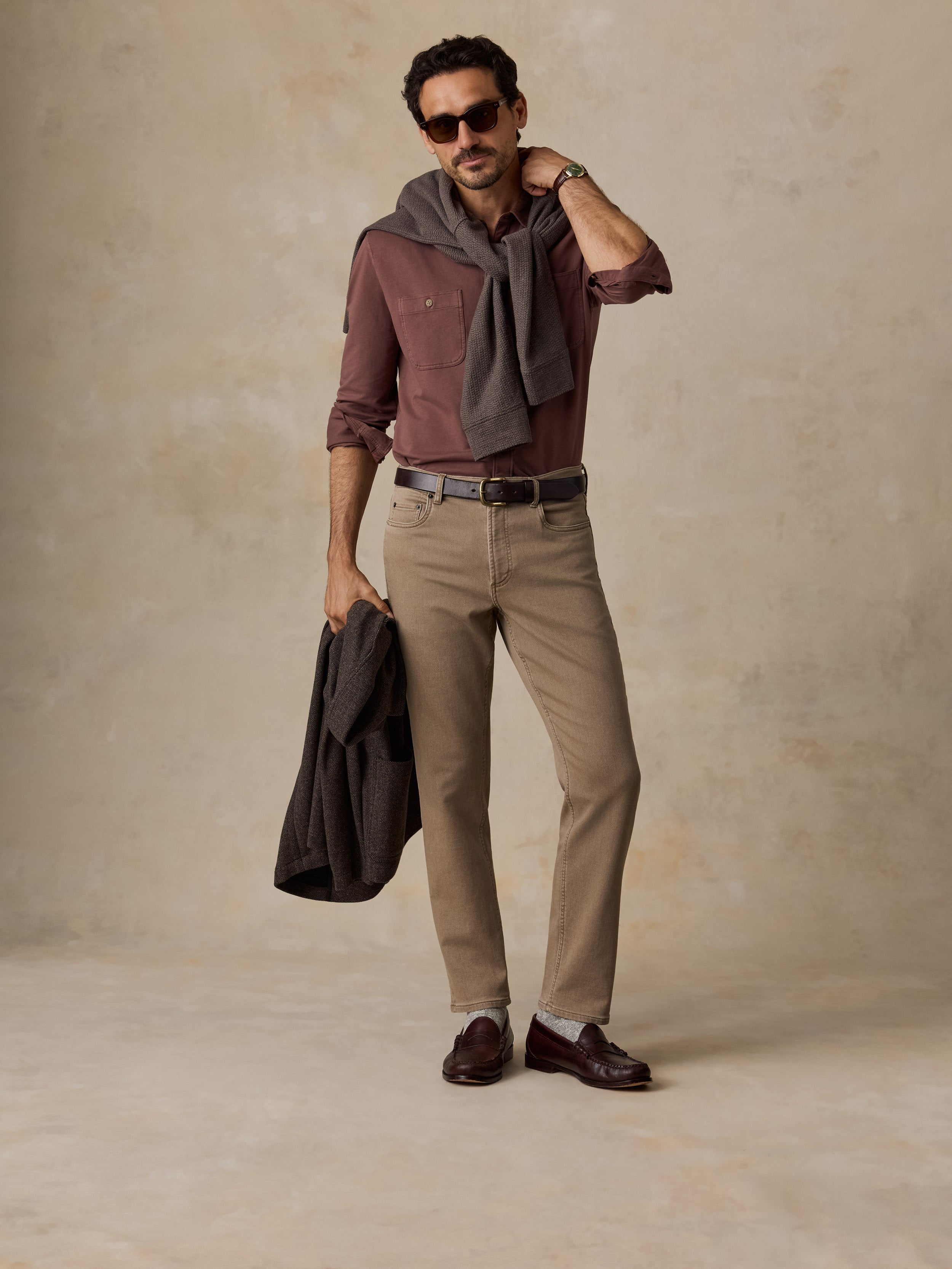 A man modeling a stylish outfit consisting of a dusty rose button-down shirt, khaki trousers, a dark gray scarf, and brown leather loafers, standing against a textured beige studio backdrop.