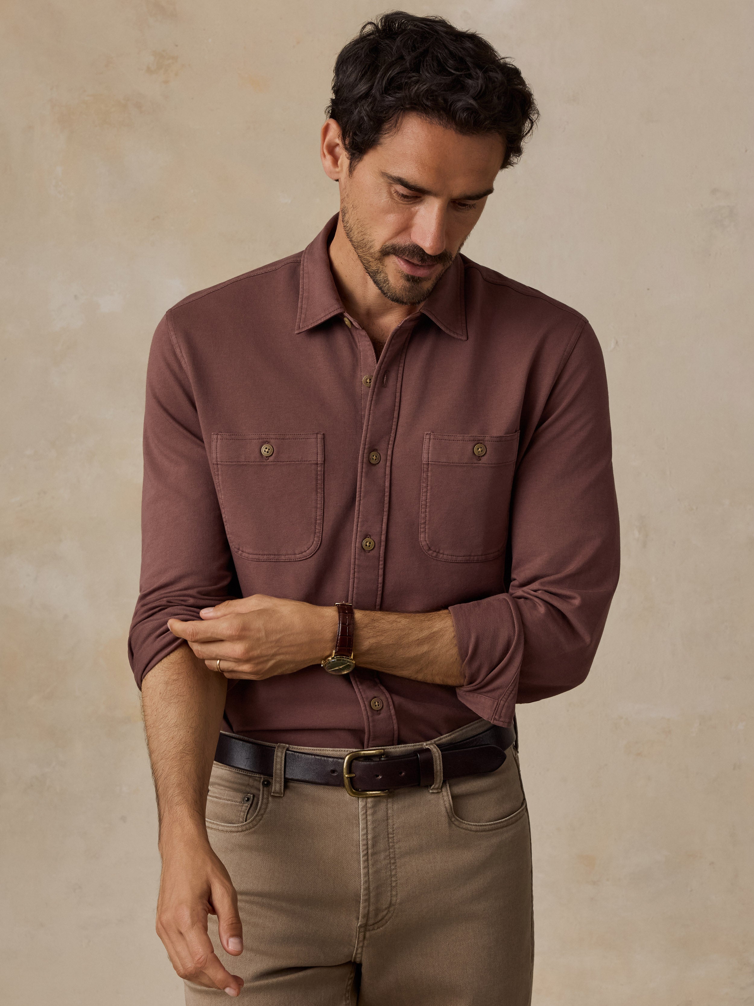 A man wearing a long-sleeve button-up shirt in a muted reddish-brown color, paired with khaki pants and a black belt, standing against a textured beige background.