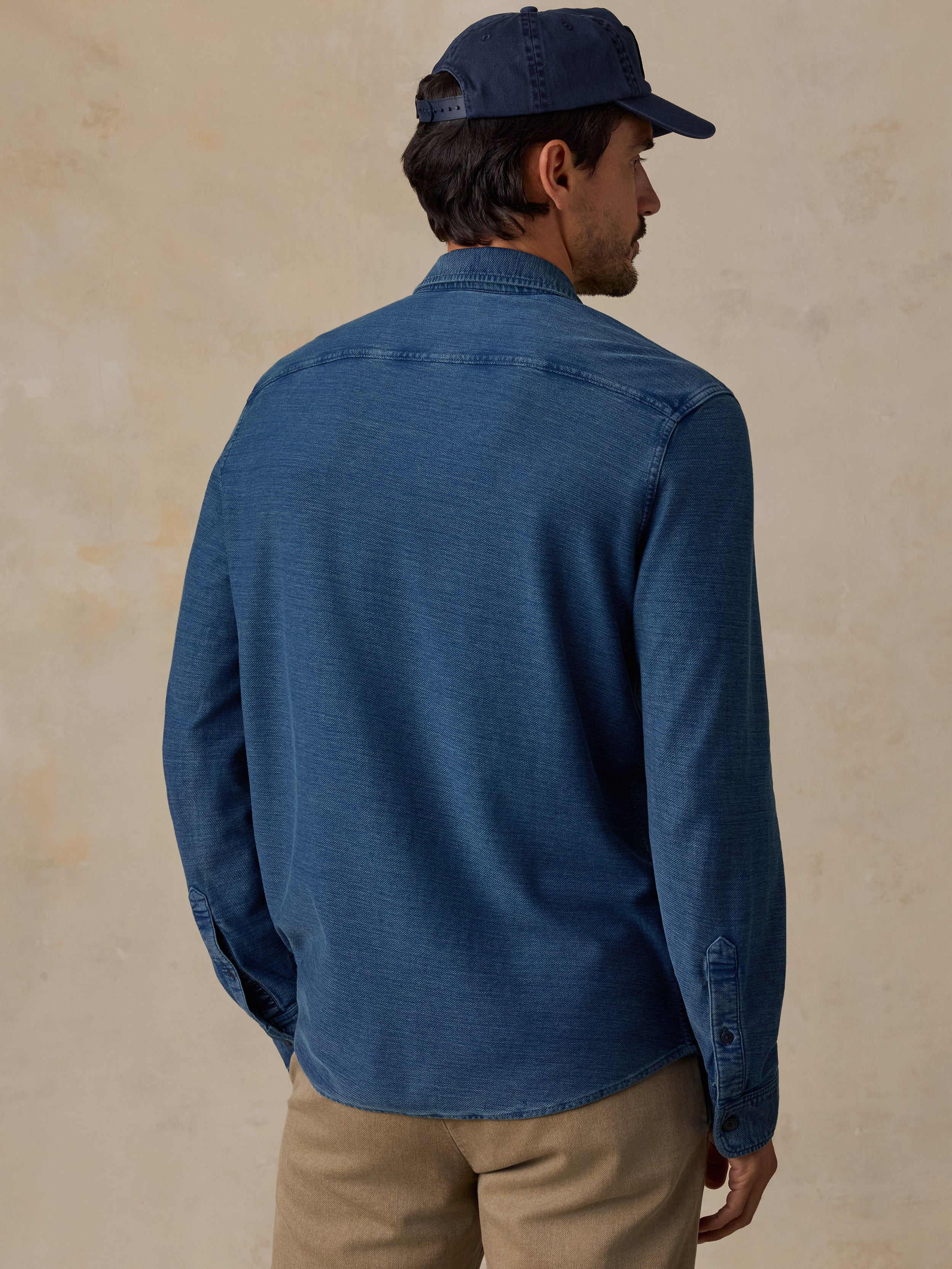 A man wearing a blue long-sleeve button-up shirt and khaki pants, shown from the back and side against a textured beige background.