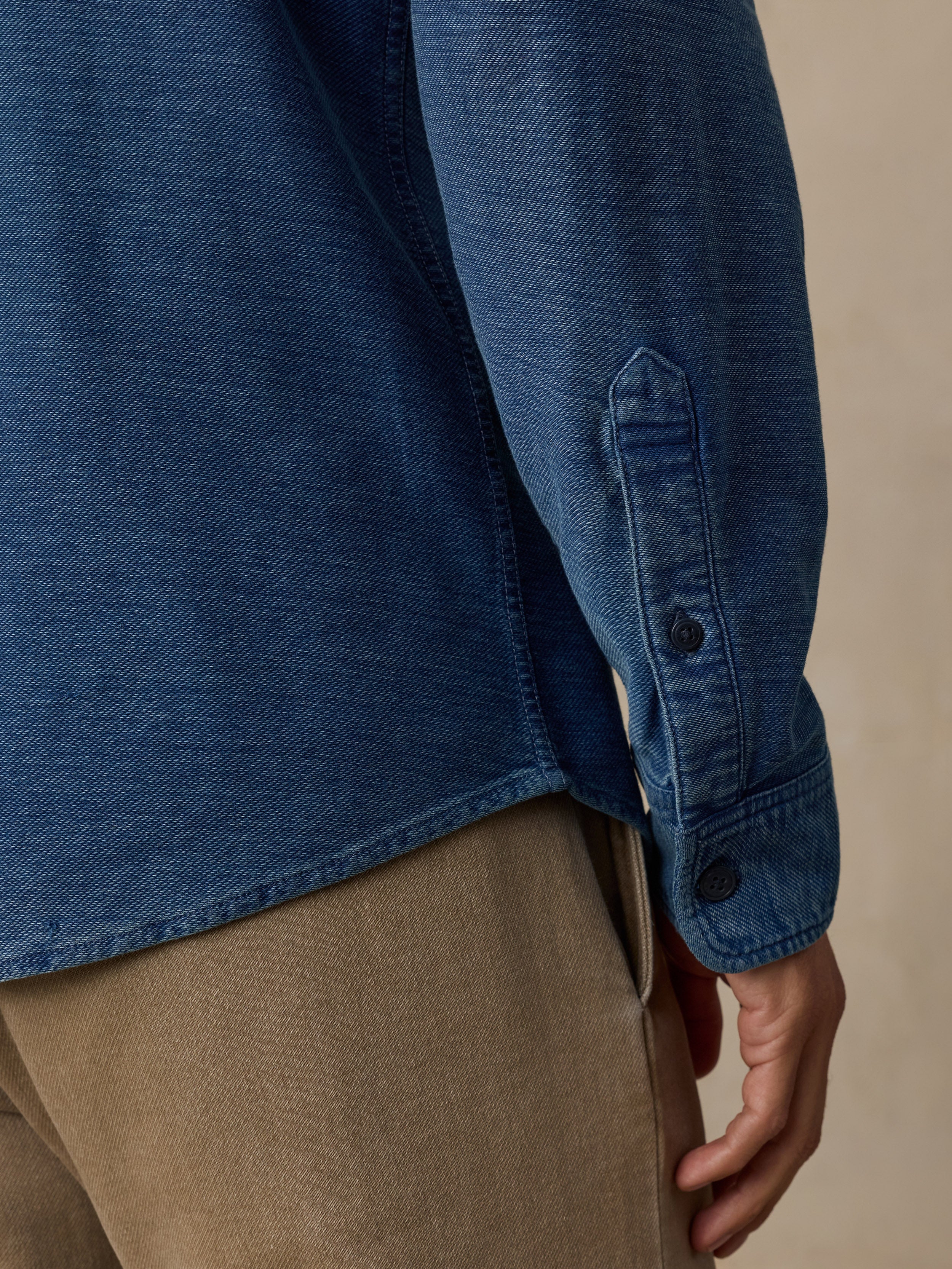 Close-up of a person wearing a blue textured long-sleeve button-up shirt and beige trousers, showing the sleeve cuff with buttons and part of the hand.