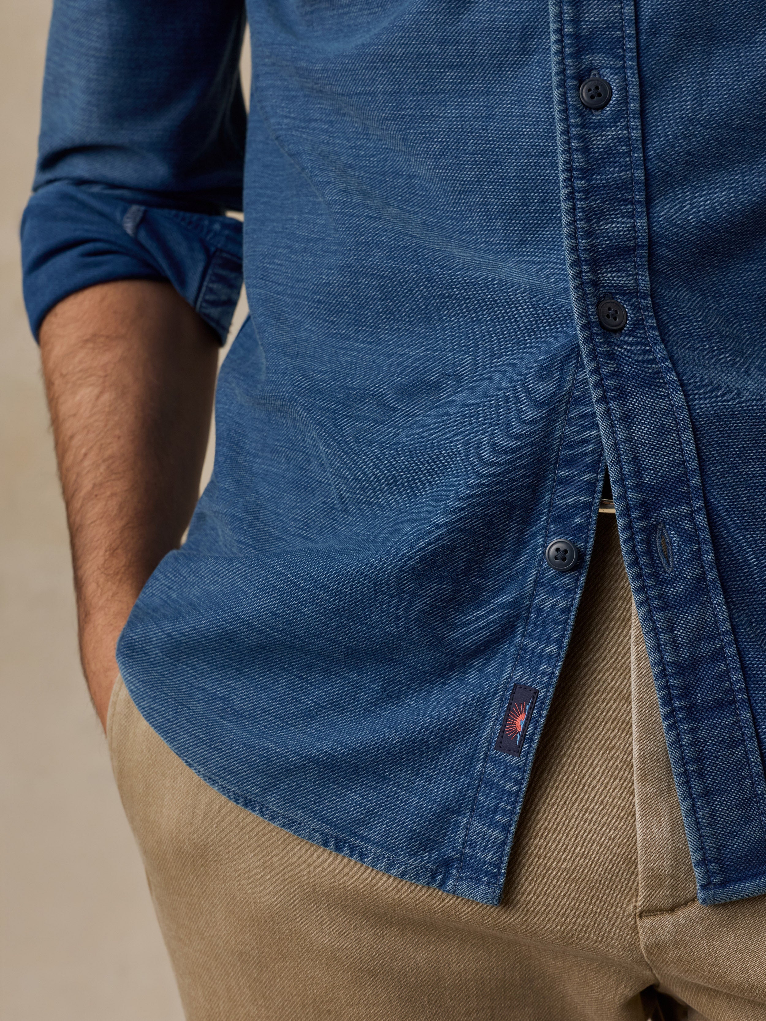 Close-up of a person wearing a blue denim button-up shirt tucked into beige trousers, with the shirt's fabric texture and a small red Faherty brand tab visible.