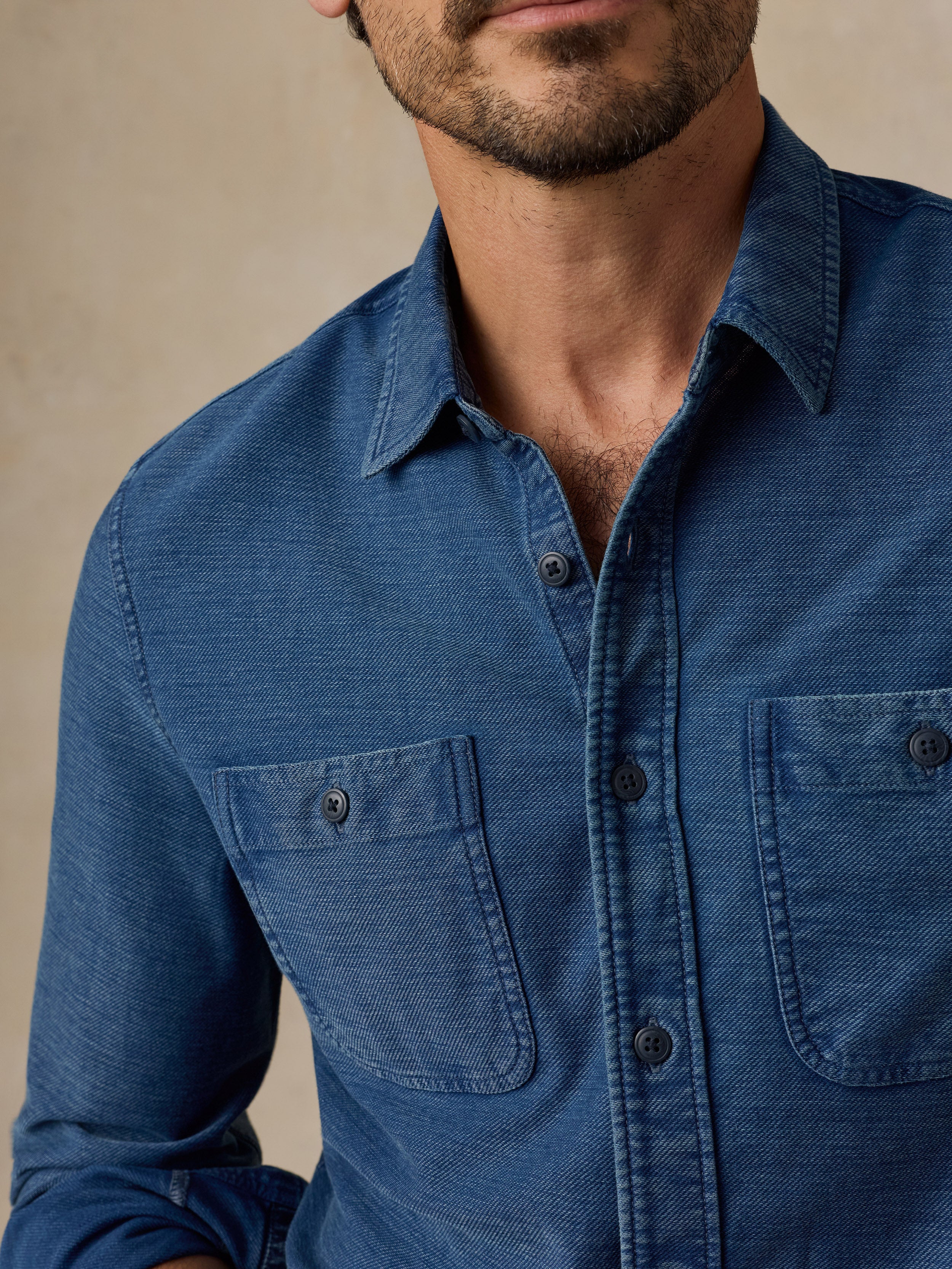A close-up of a man wearing a textured blue long-sleeve button-up shirt with two chest pockets, set against a neutral beige background.