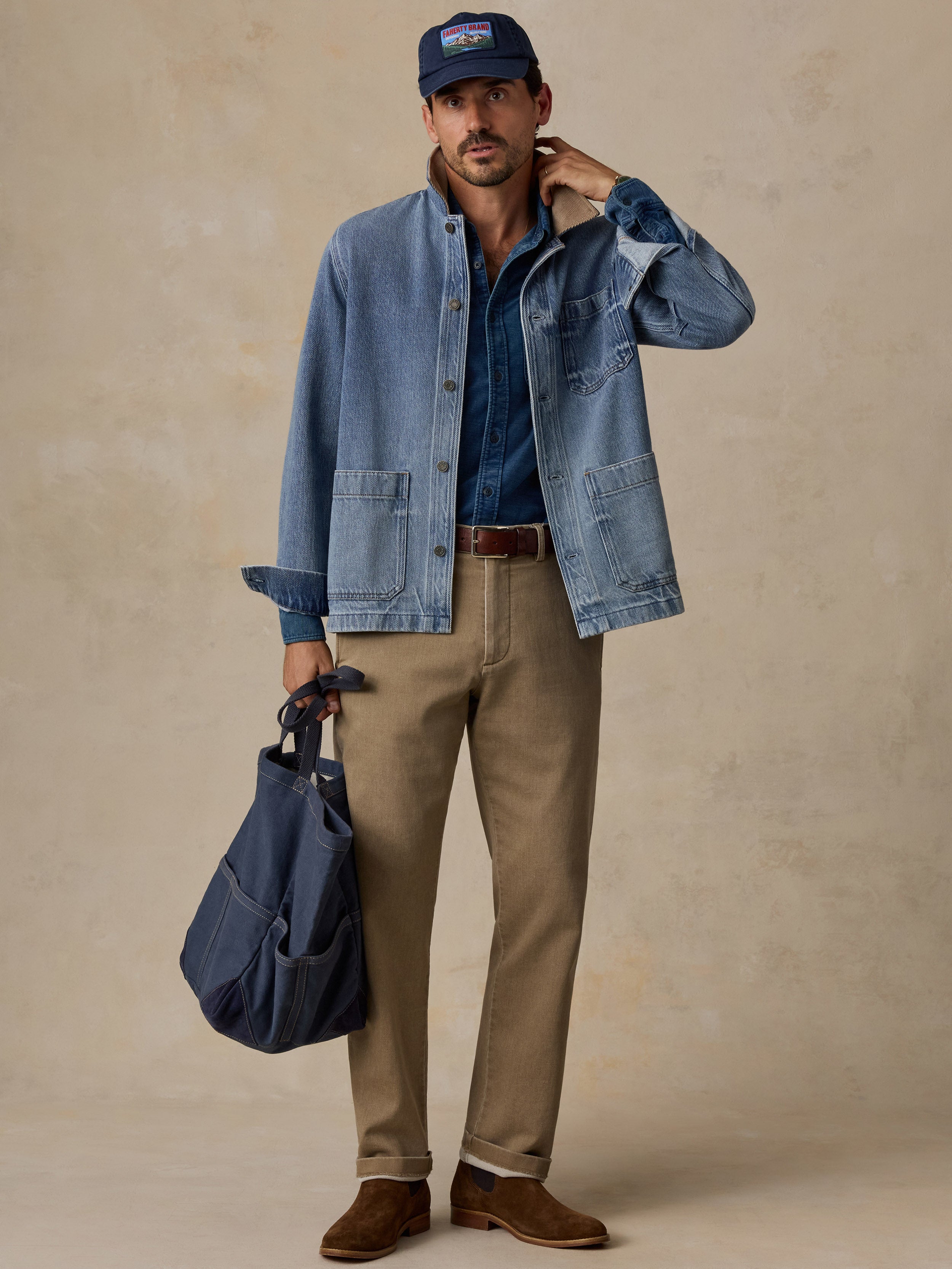 A man models a light blue denim jacket over a dark blue button-down shirt, paired with khaki pants, brown suede shoes, a navy cap, and a navy tote bag, set against a textured beige studio backdrop.