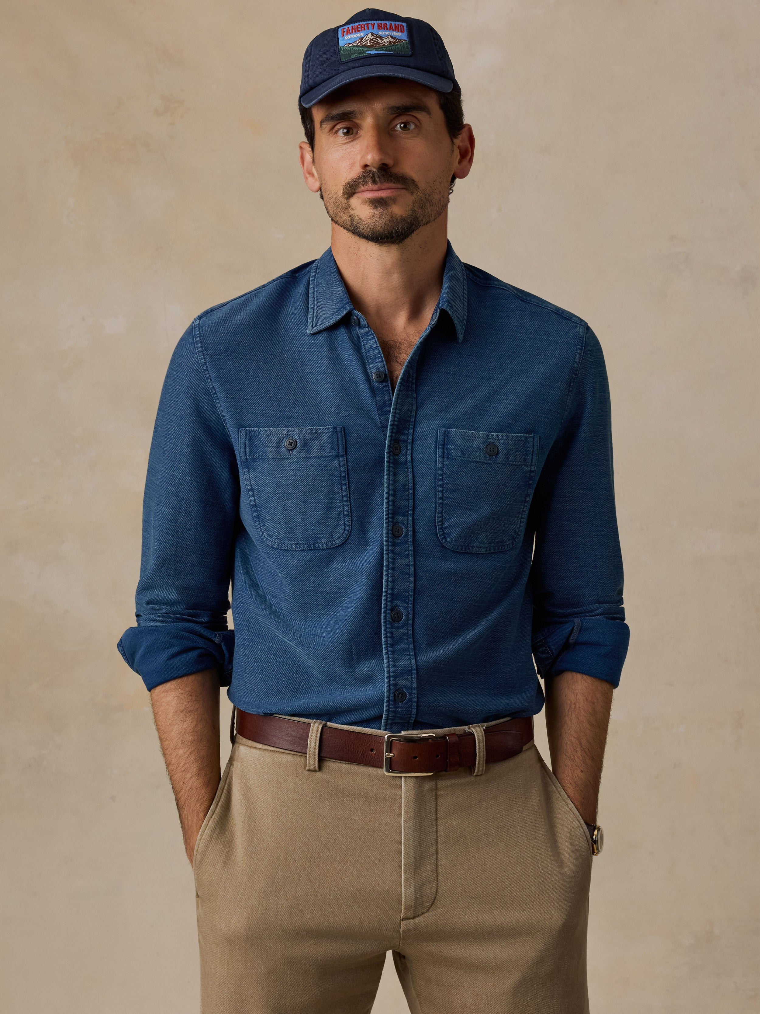 A man wearing a blue long-sleeve button-up shirt with two chest pockets, khaki pants, a brown belt, and a navy baseball cap, standing against a textured beige background.