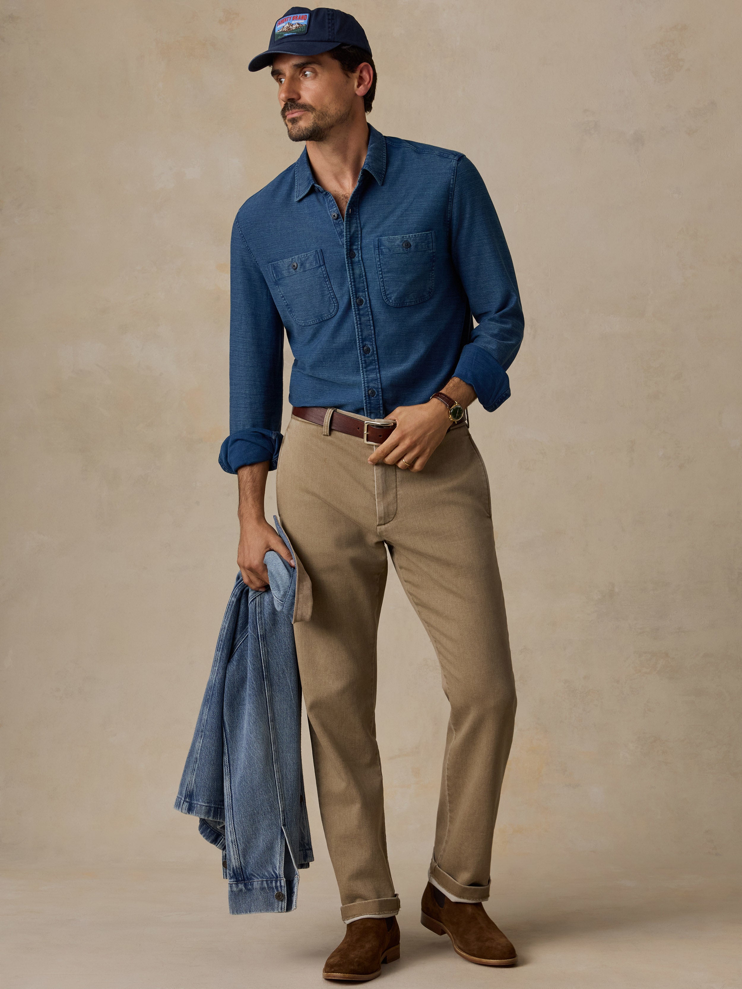 A man stands against a textured beige studio backdrop wearing a blue long-sleeve button-up shirt, tan chinos, brown suede shoes, and a navy baseball cap, while holding a folded blue denim jacket.