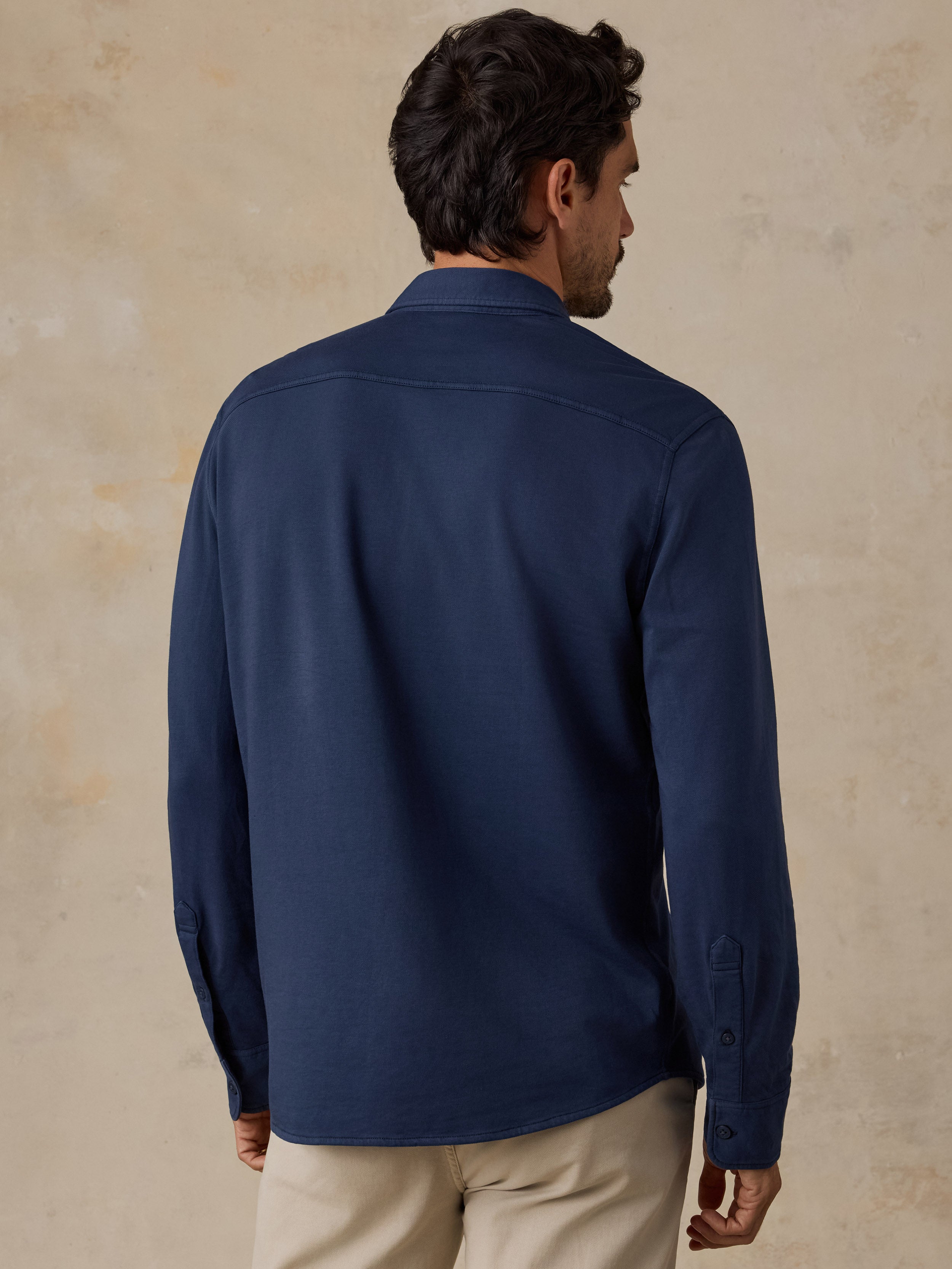 A man viewed from the back wearing a navy blue long-sleeve button-up shirt and beige trousers, standing against a textured beige wall.