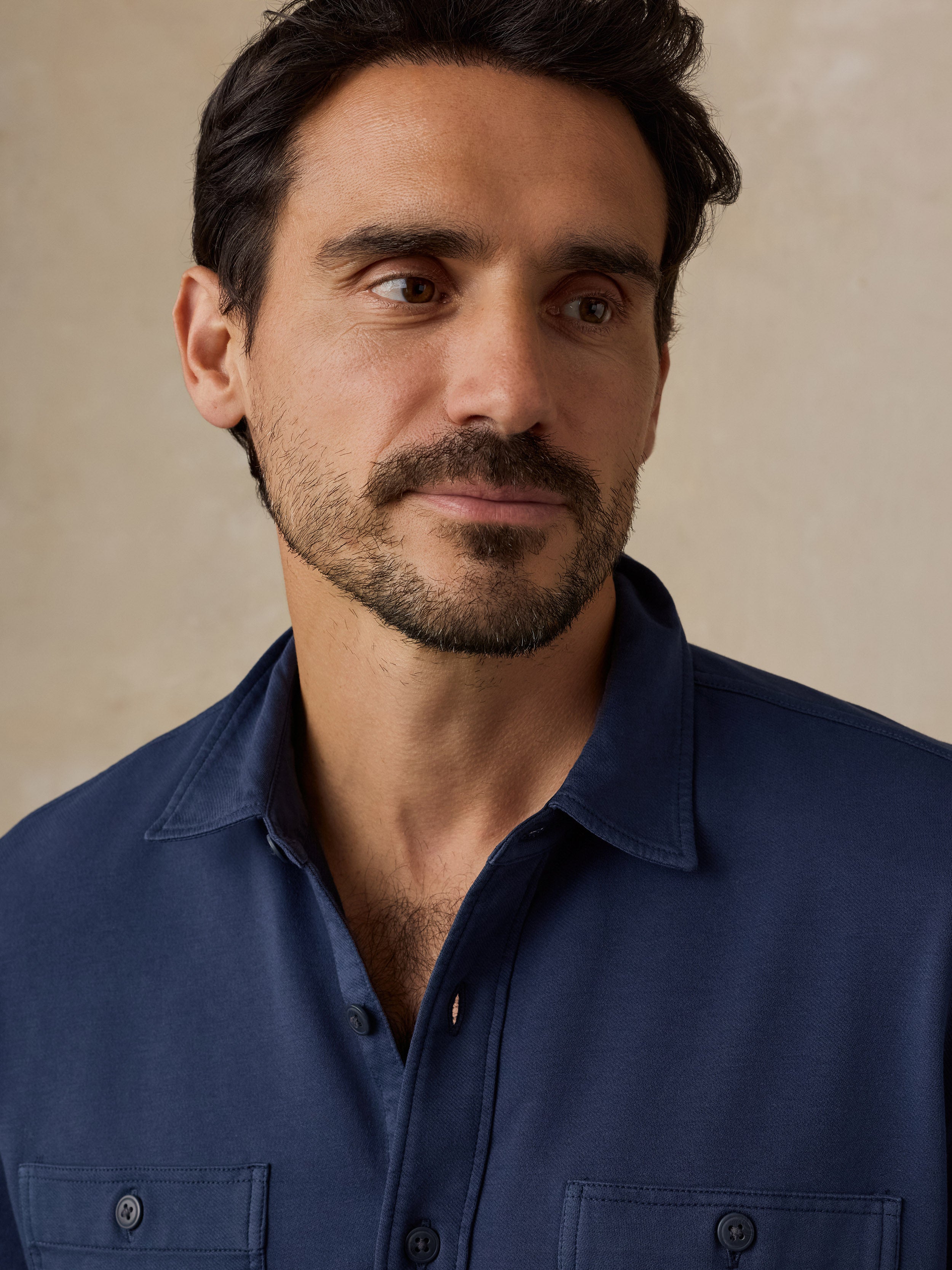A close-up portrait of a man wearing a dark navy blue button-down shirt against a neutral beige background.