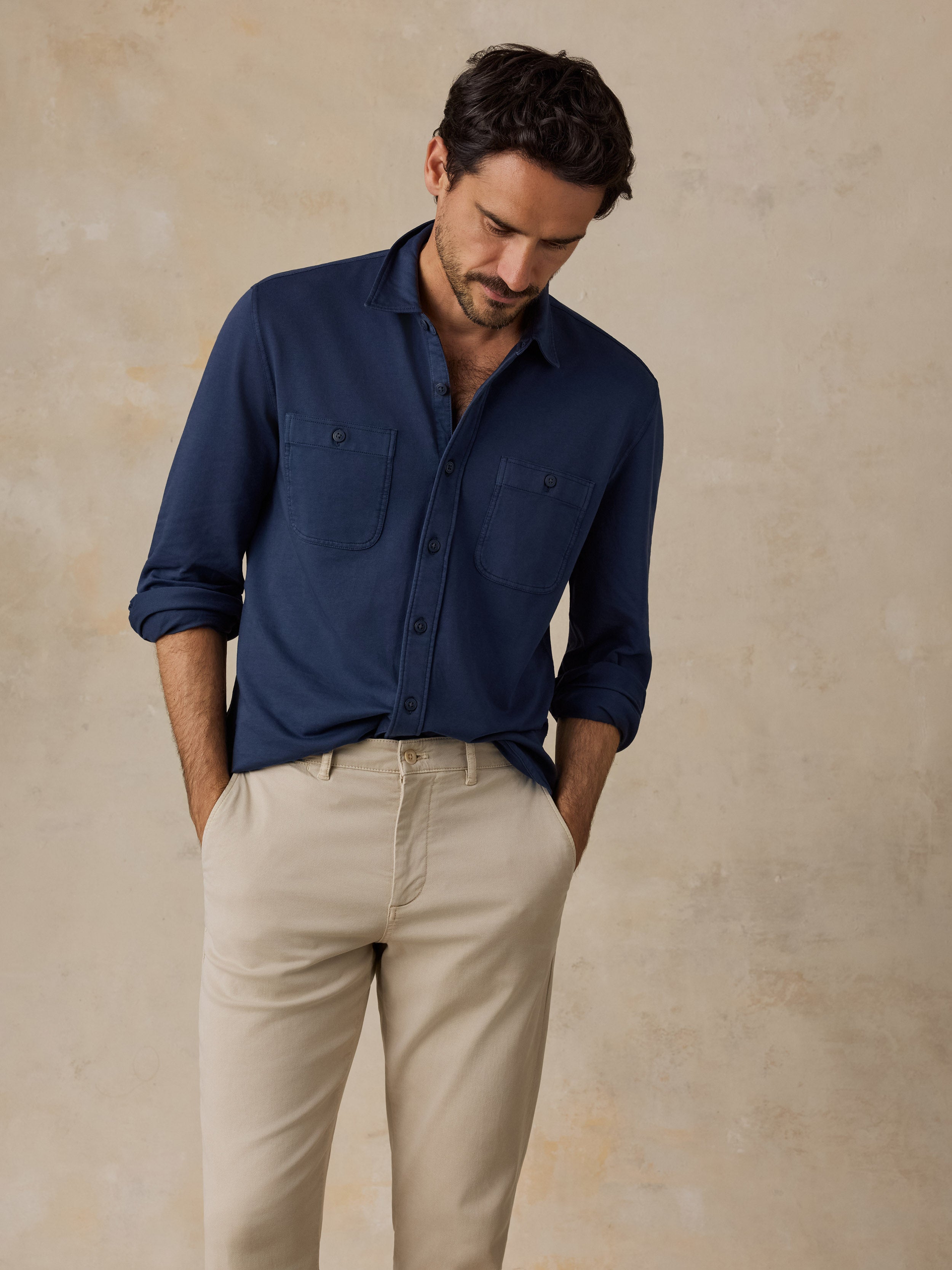 A man wearing a navy blue long-sleeve button-up shirt with two chest pockets and beige trousers, standing against a textured beige wall.