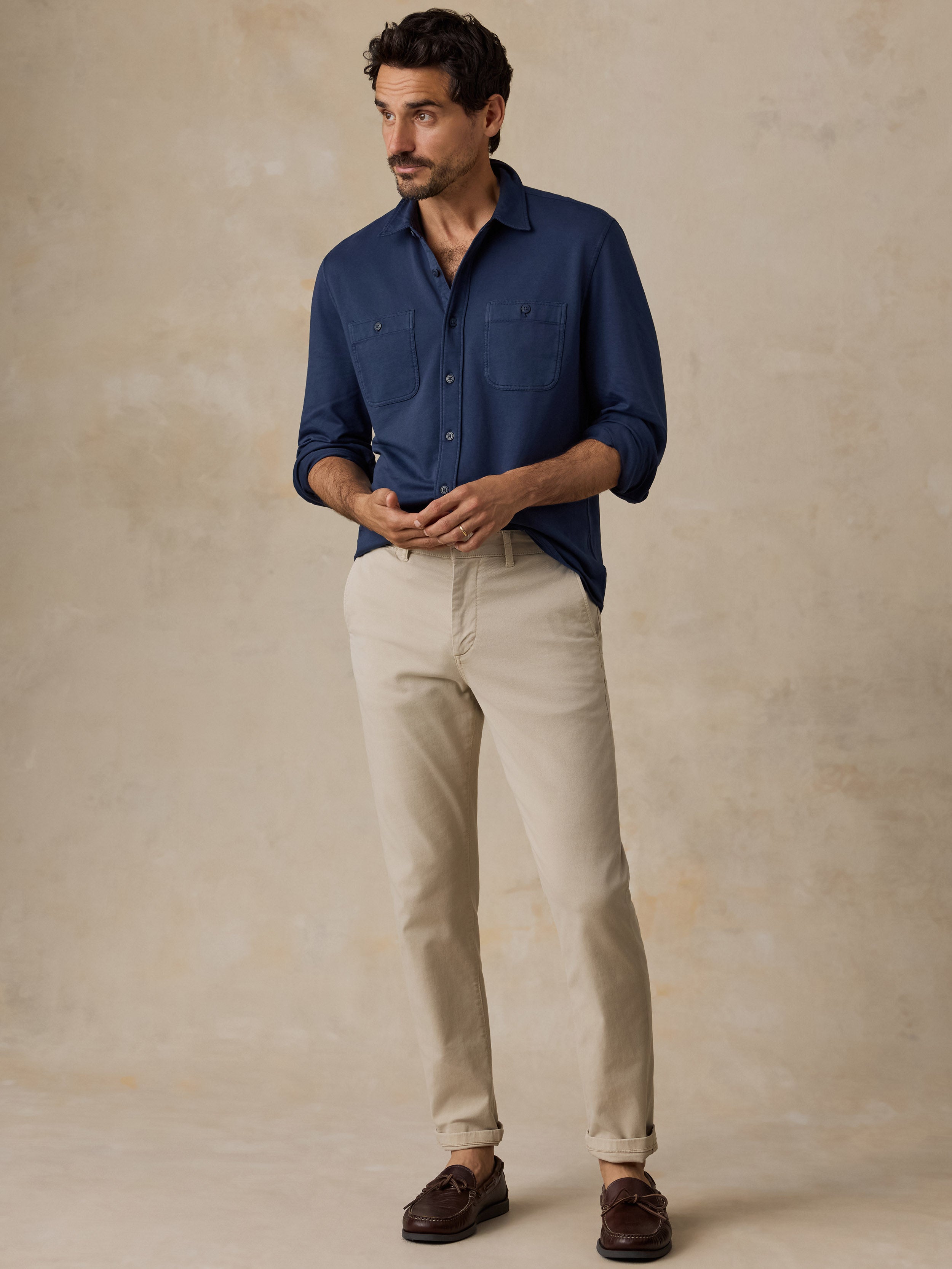 A man modeling a navy blue long-sleeve button-down shirt and beige trousers, standing against a textured beige studio backdrop.