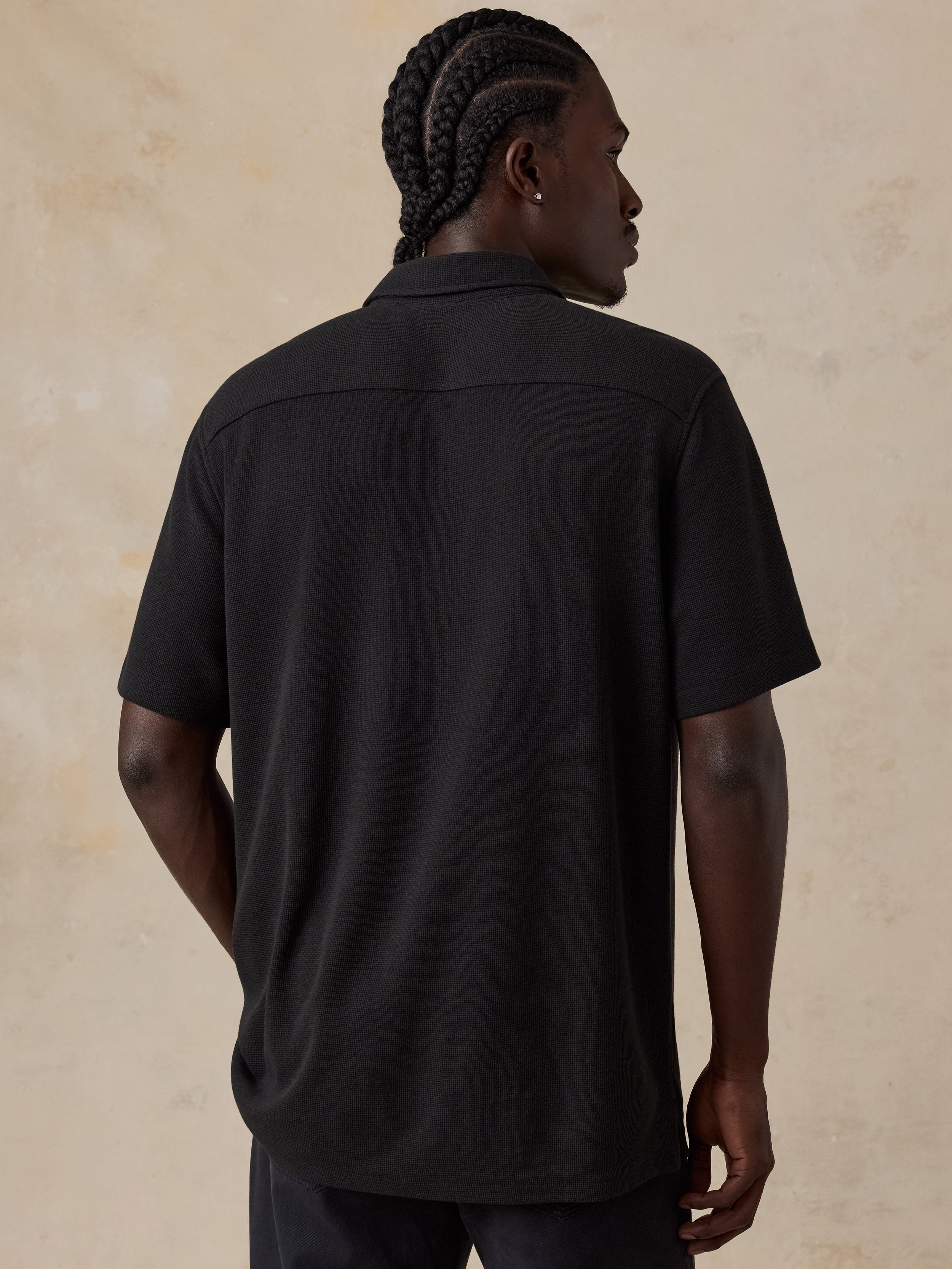 A man with braided hair is shown from the back wearing a black short-sleeve collared shirt against a textured beige background.