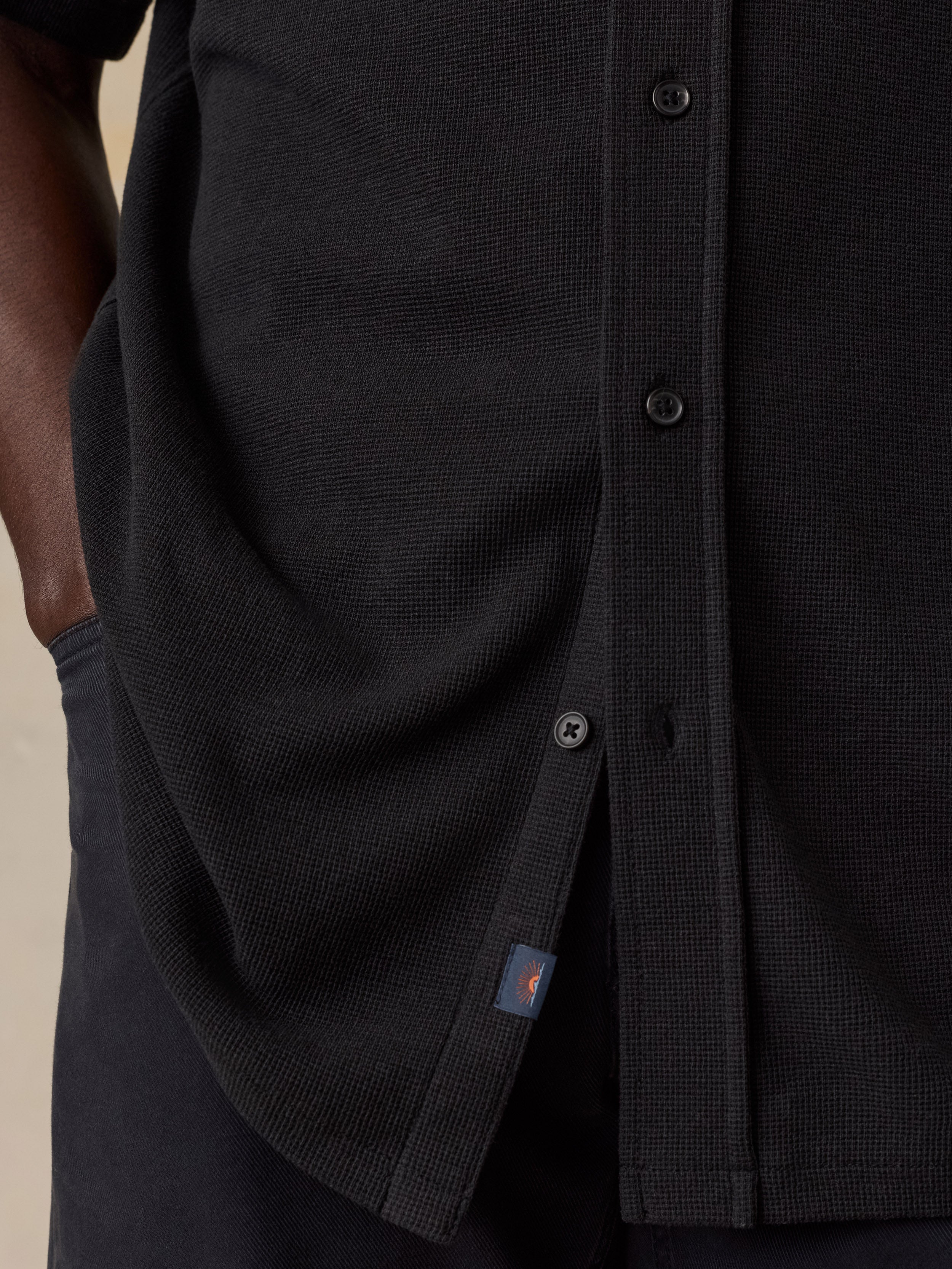 Close-up of a black textured button-up shirt worn by a model, showing the ribbed fabric, dark buttons, and a small orange and blue Faherty brand tag near the hem.