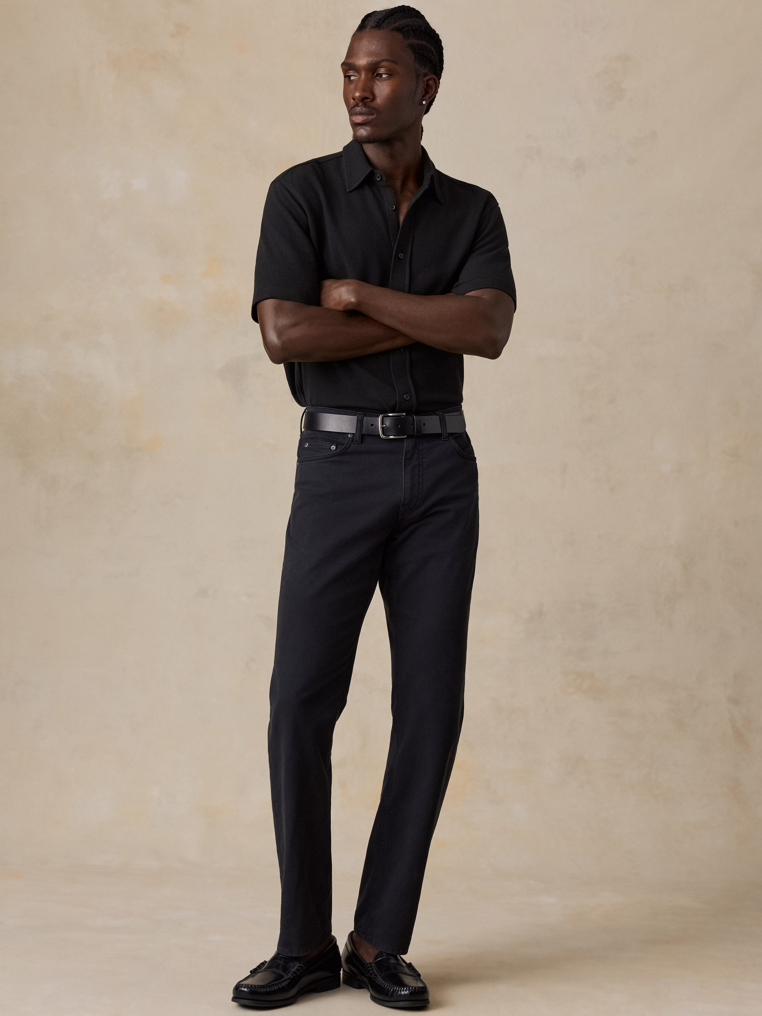A man modeling a black short-sleeve button-up shirt and black trousers with a black belt, standing against a textured beige studio backdrop.