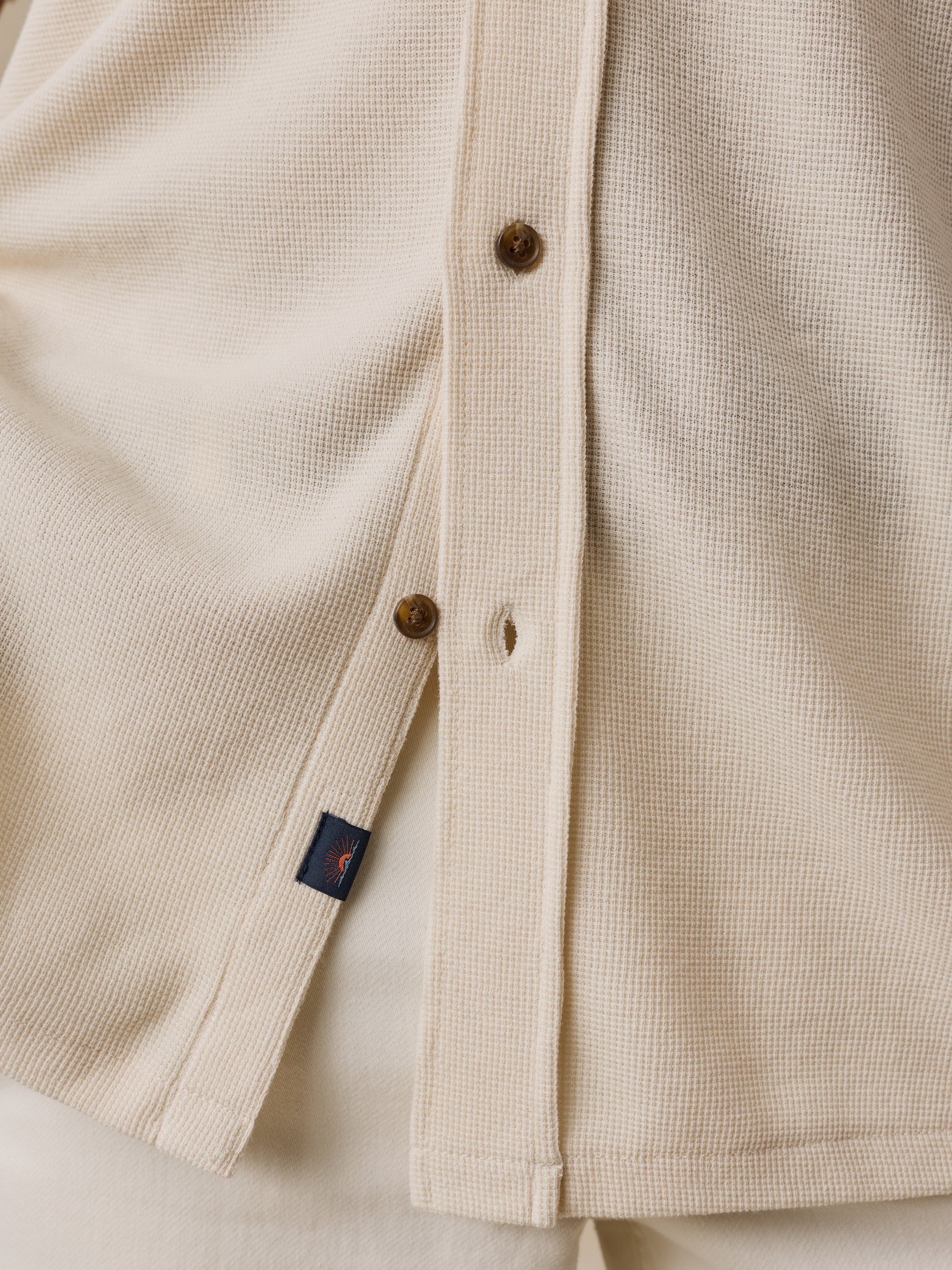 Close-up of a cream-colored waffle-knit button-up shirt with brown buttons and a small navy Faherty brand tag.