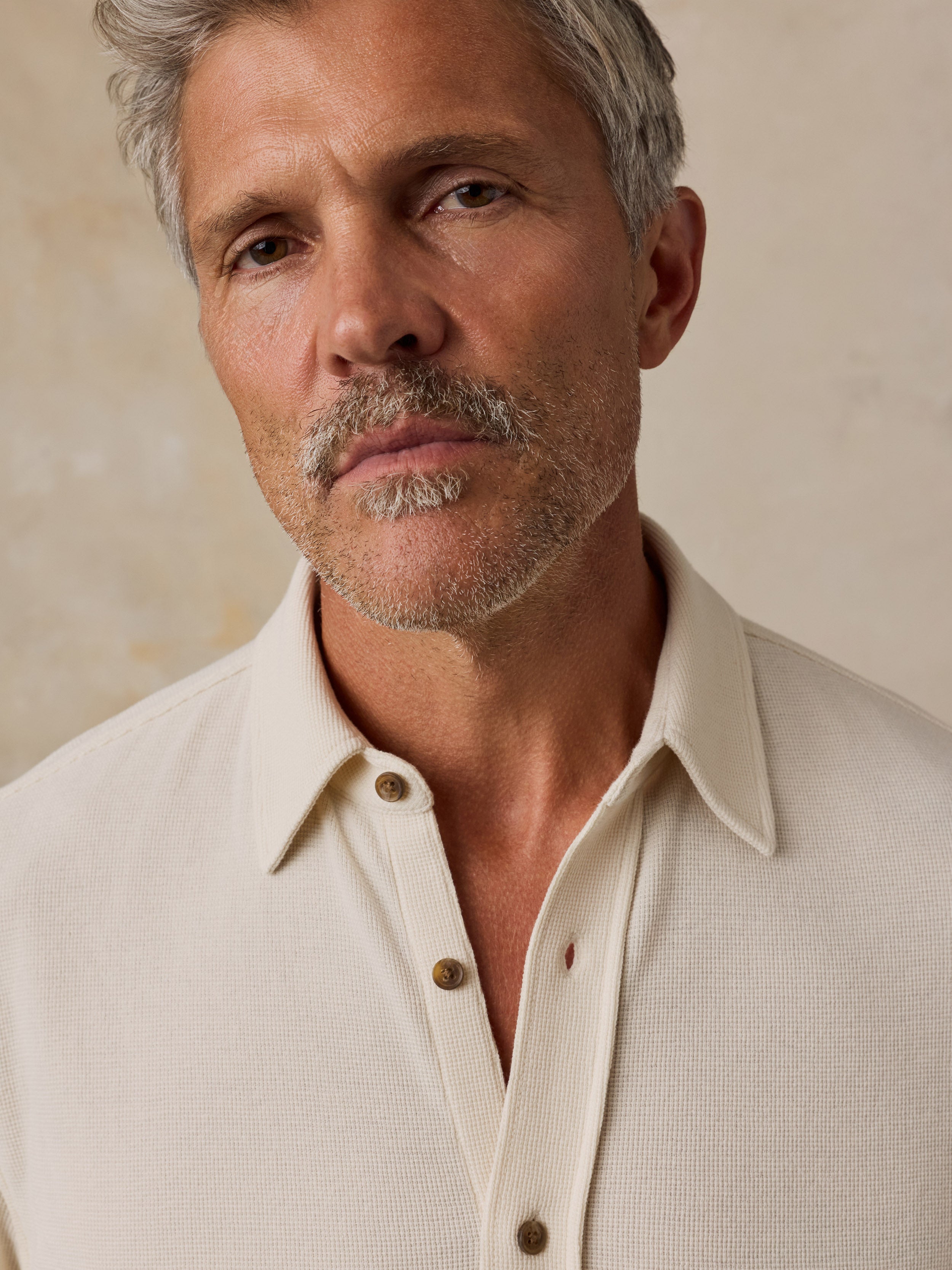 A close-up portrait of a man with gray hair and a short beard, wearing a cream-colored textured knit polo shirt against a neutral beige background.