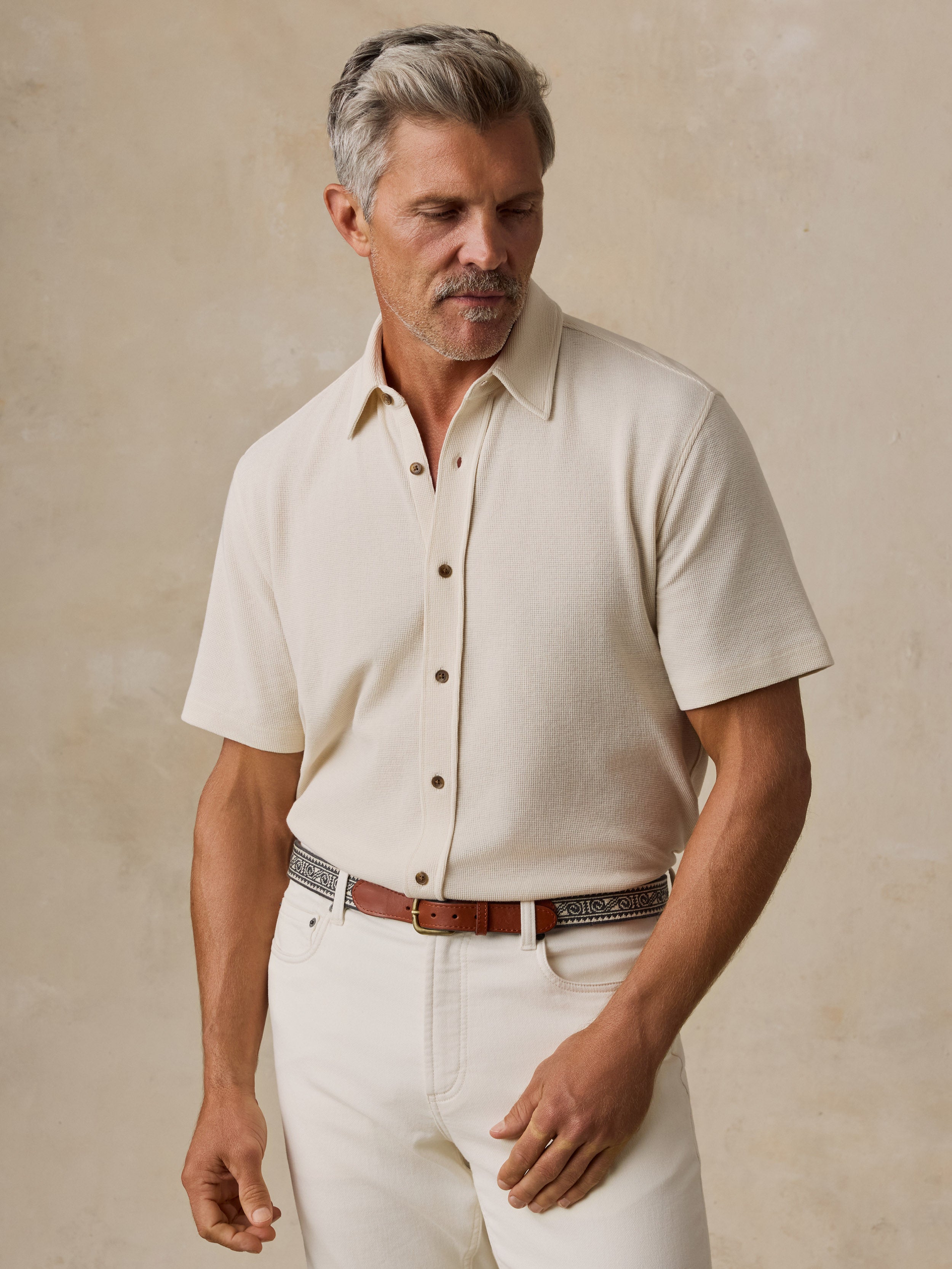 A man wearing a short-sleeve cream-colored knit button-up shirt and white pants, standing against a textured beige background.