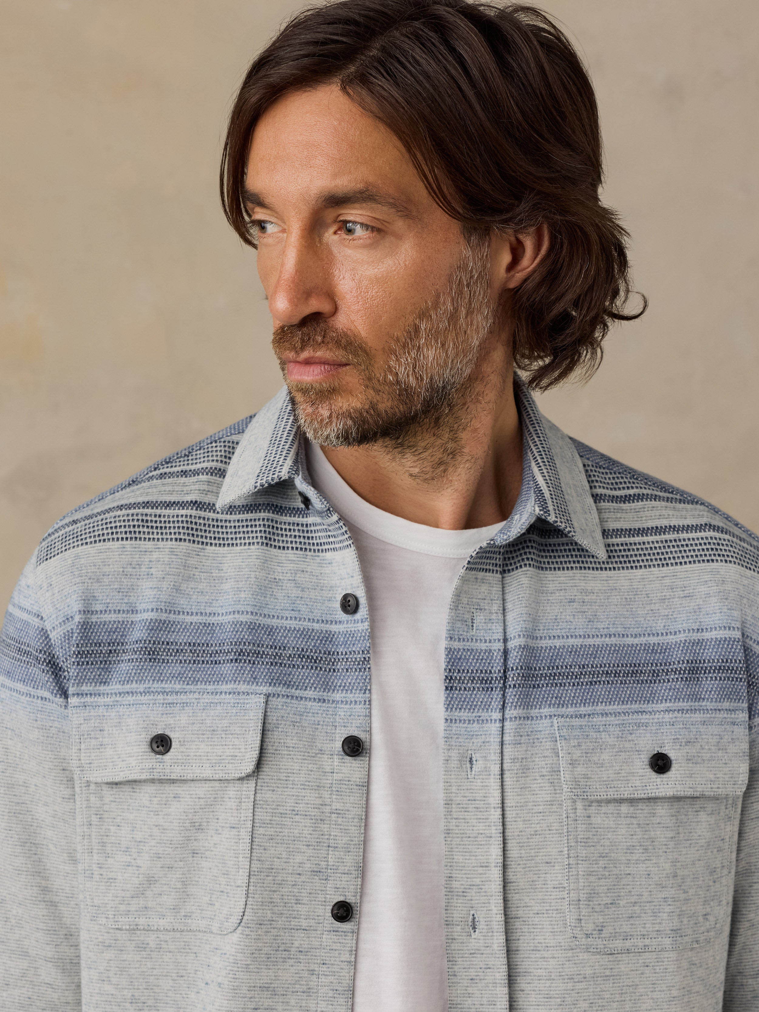A man wearing a light gray and blue striped button-down shirt over a white t-shirt, photographed against a neutral beige background.