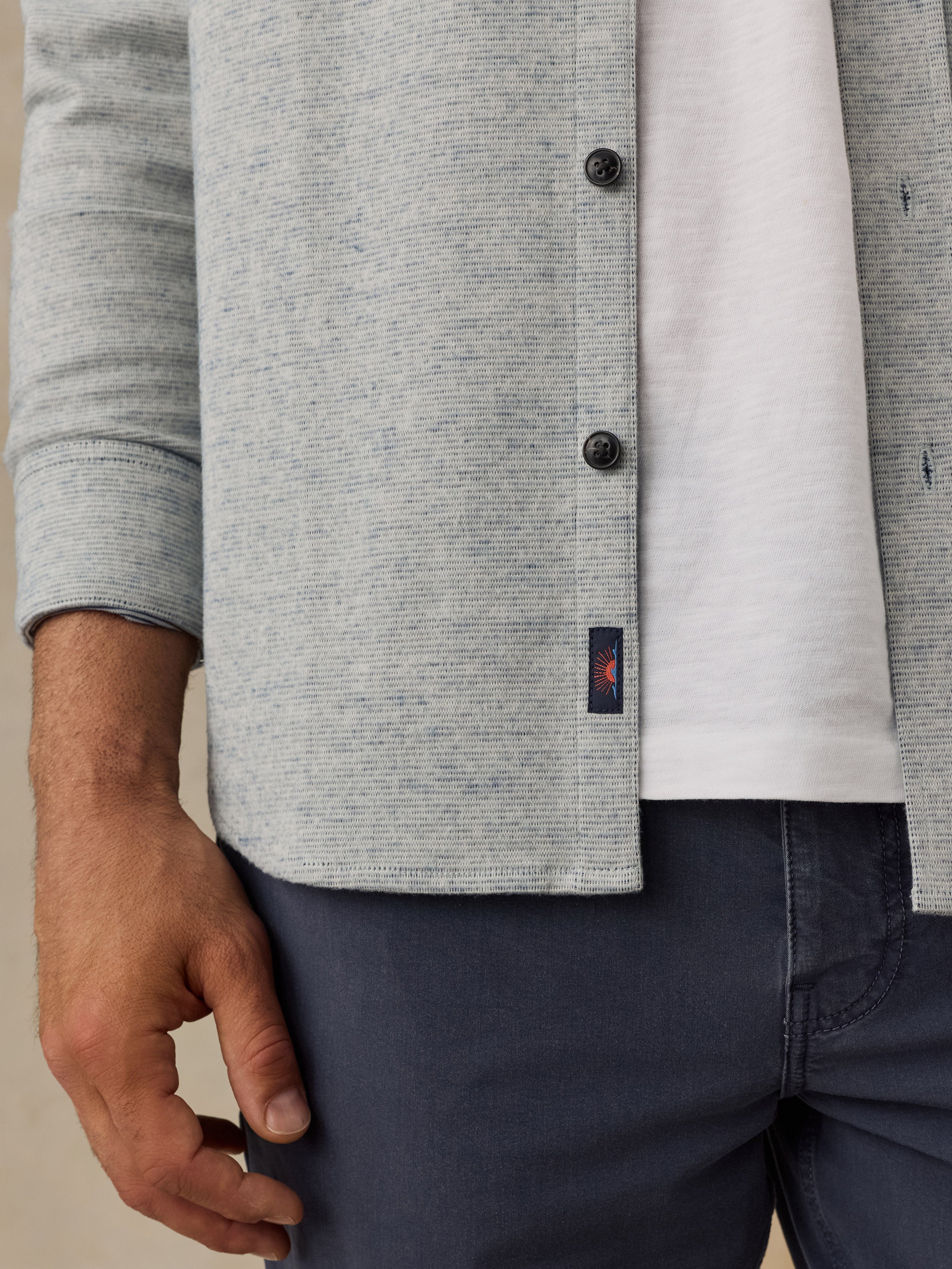 Close-up of a man wearing a light gray heathered knit cardigan over a white T-shirt and navy pants, with a small Faherty brand tag visible near the hem.