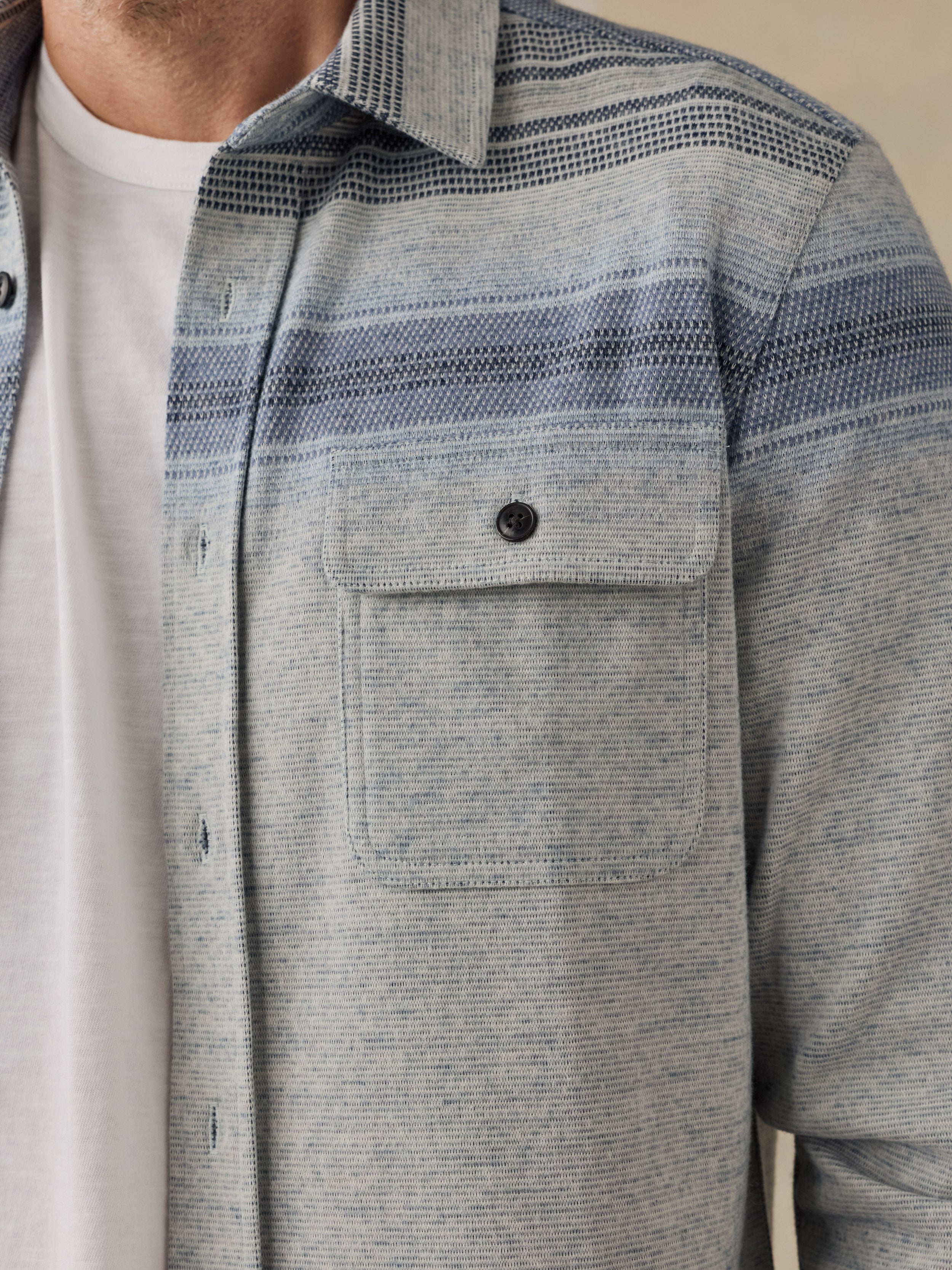 Close-up of a man wearing a light gray and blue striped textured button-down shirt over a white t-shirt.