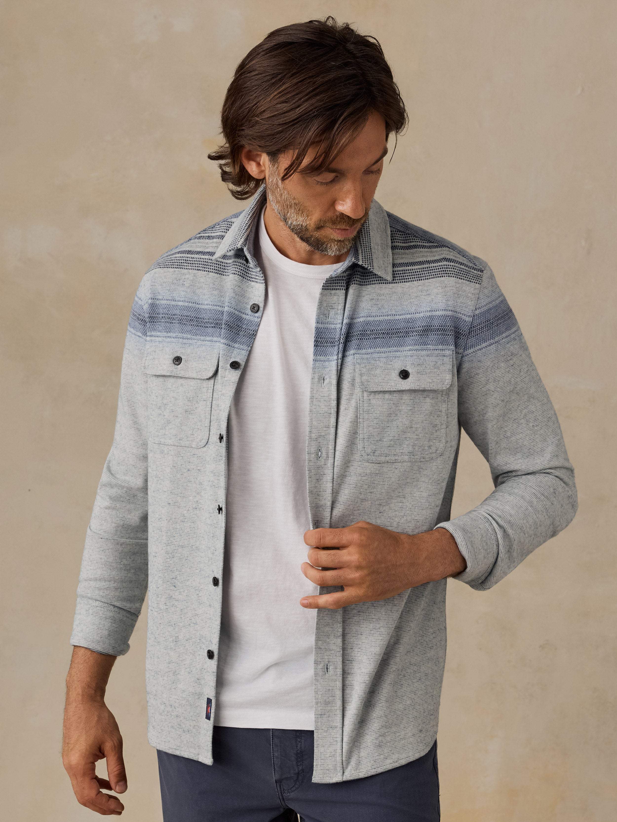 A man wearing a light gray heathered button-up shirt with blue and gray horizontal stripes across the chest, layered over a white t-shirt and dark blue jeans, standing against a textured beige background.