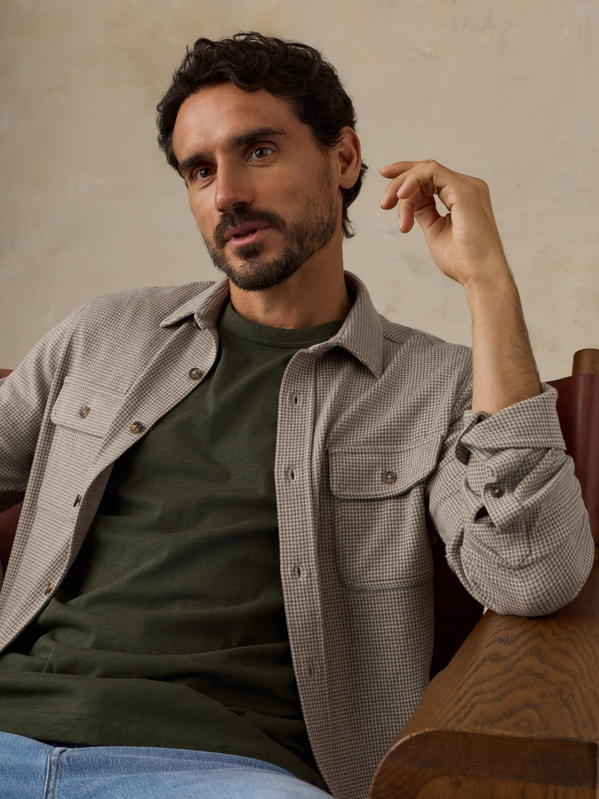 Man sitting on a wooden chair wearing a beige overshirt with a green t-shirt and jeans against a beige studio background, image 2 of 7