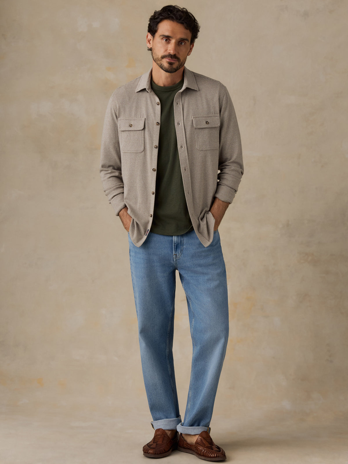 Man wearing a beige overshirt with a green t-shirt and jeans against a beige studio background, image 7 of 7
